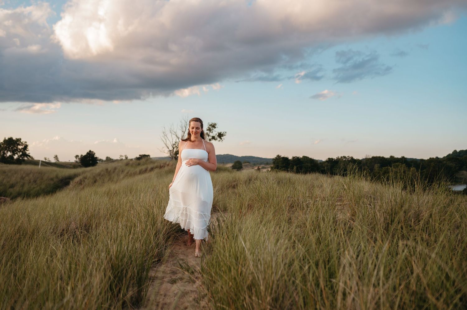 A figure in a flowing white dress walks along a grassy hilltop path at sunset with clouds in the sky.