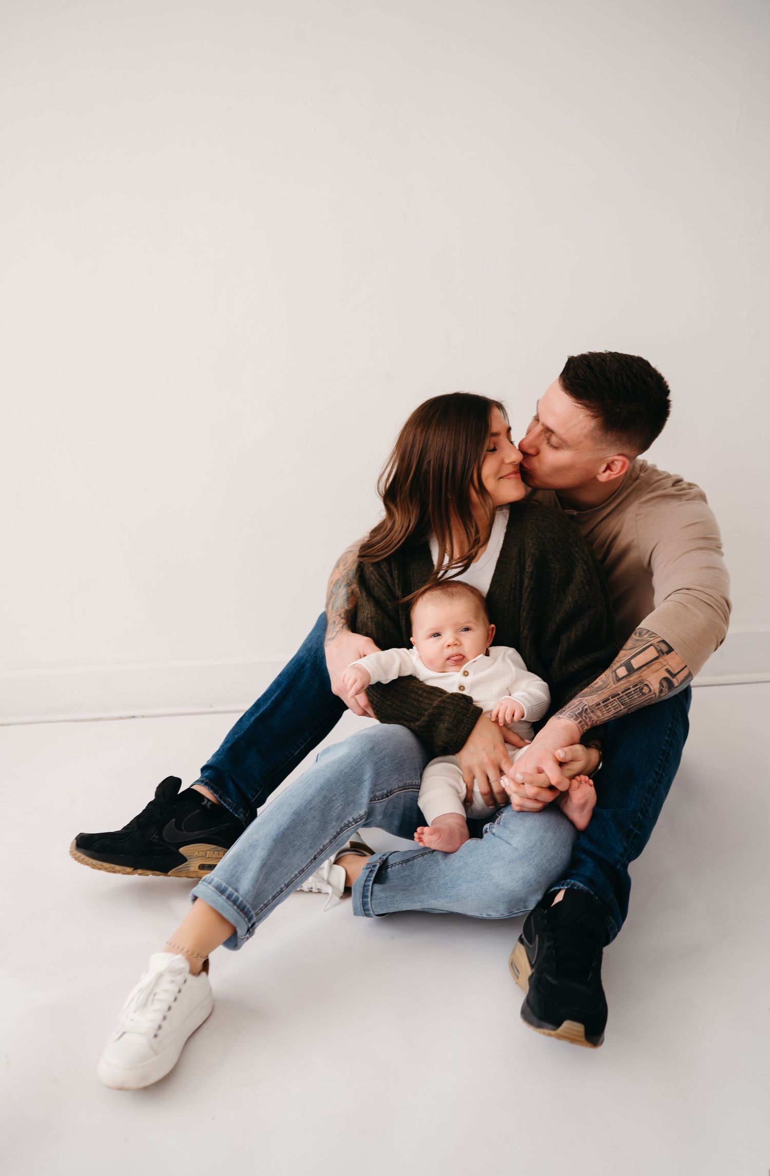 A couple sits with their baby in casual clothing against a white backdrop while sharing an intimate moment together.