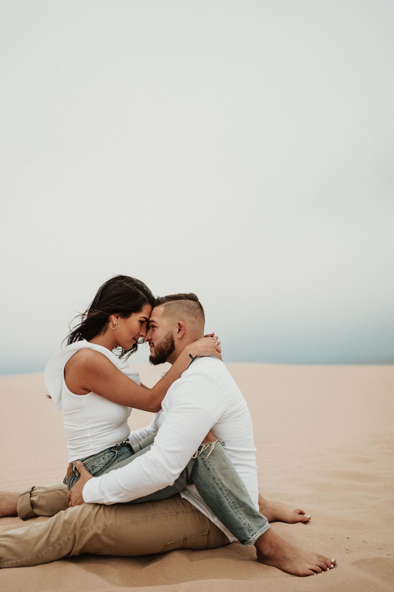 A couple embraces tenderly while sitting on sandy dunes during sunset.