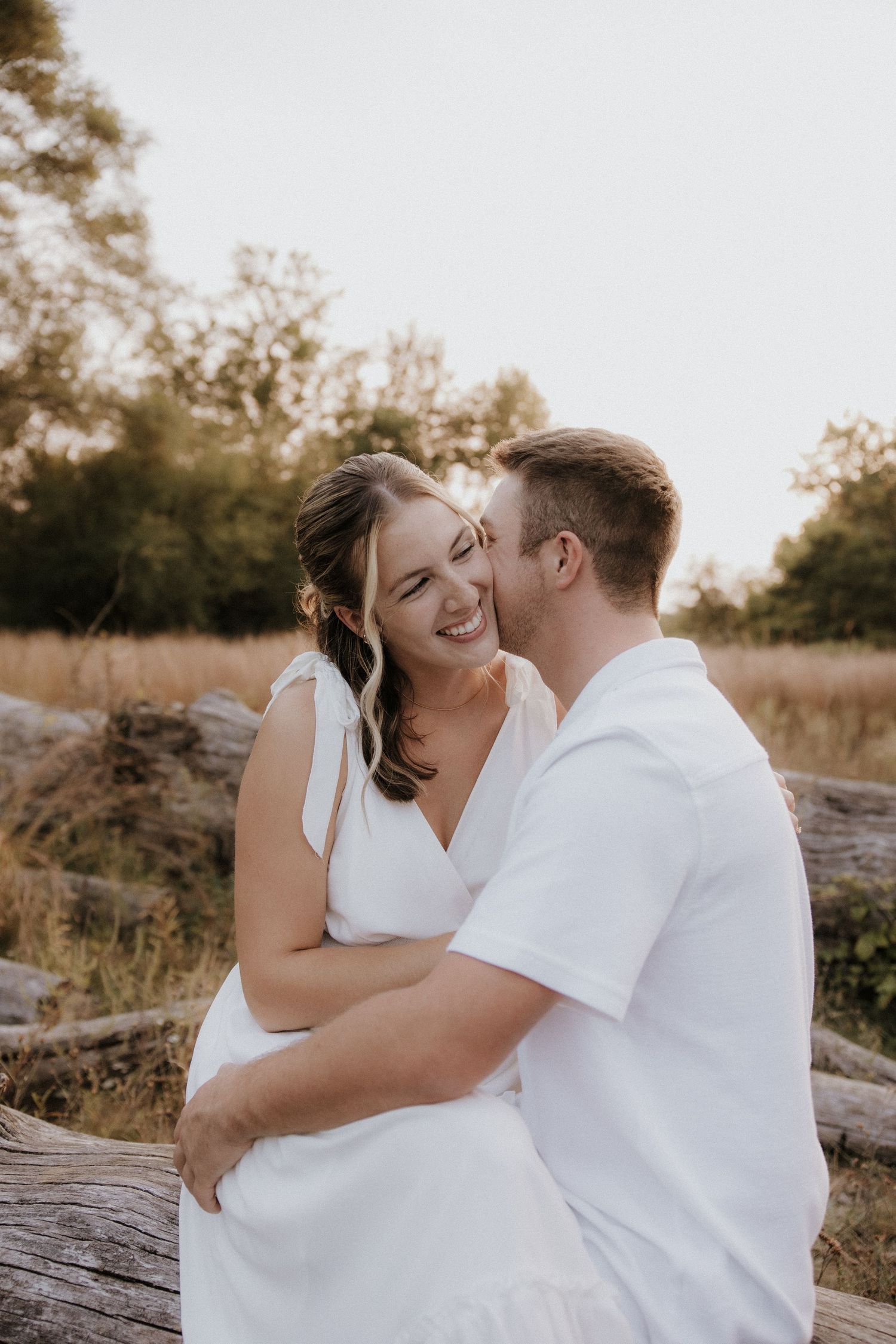 Romantic couple in white clothing embracing on fallen tree during golden hour photoshoot.