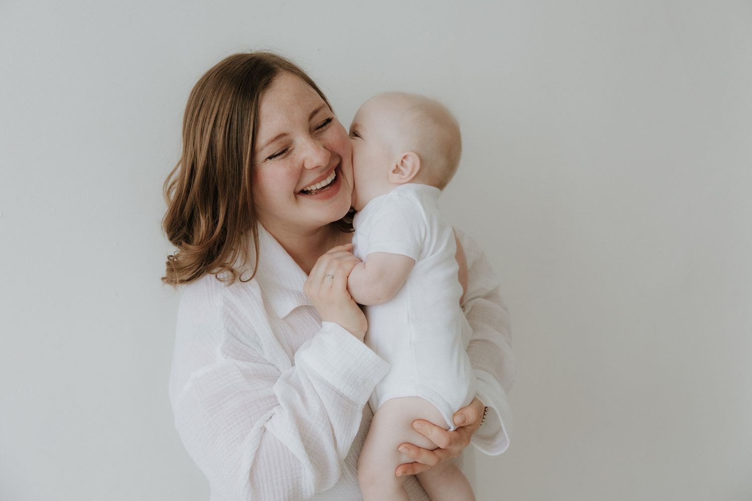 Happy laughing mother in white shirt holding smiling baby against light background.