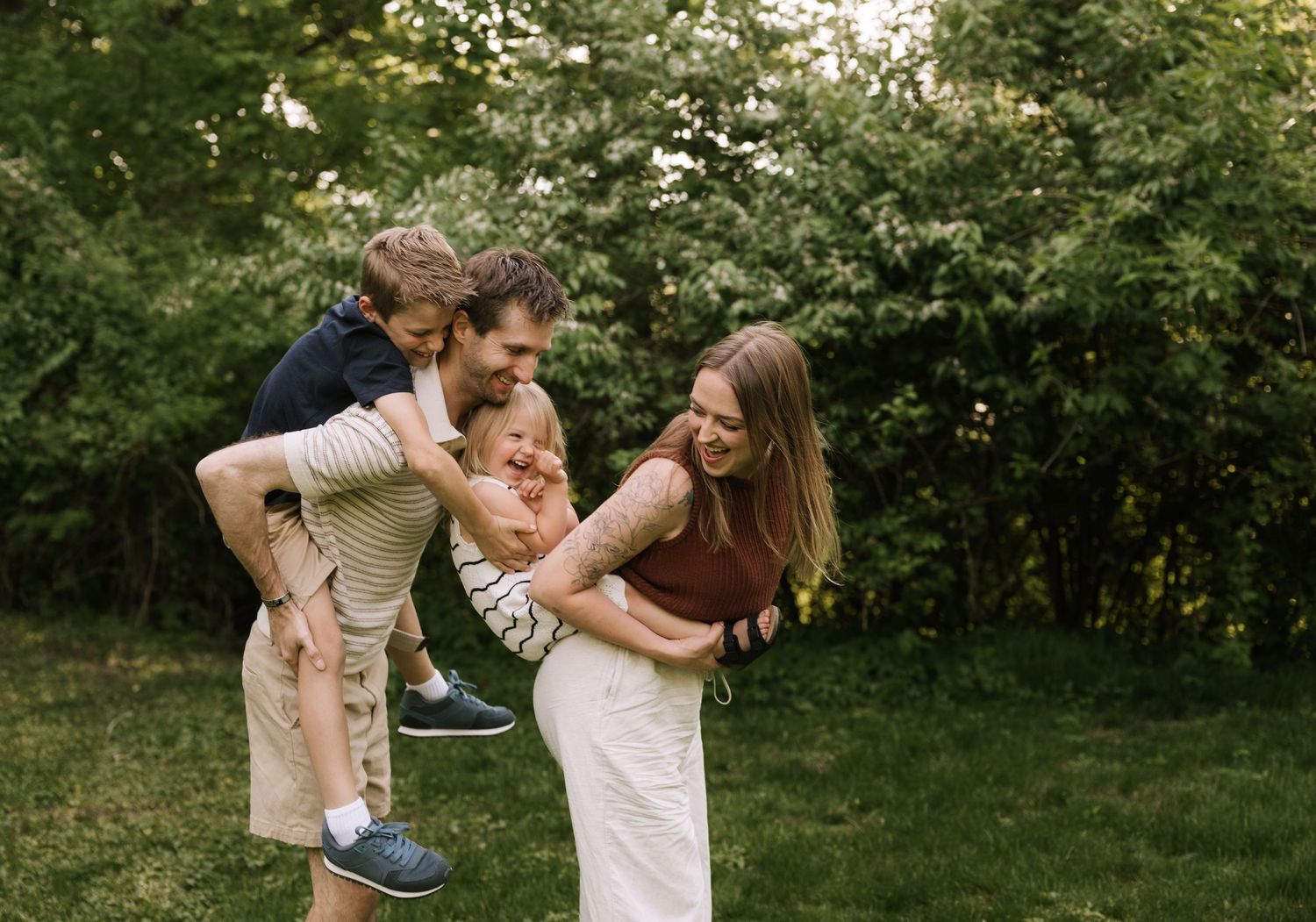 A joyful family plays outdoors in a lush green garden, with parents laughing as they hold their young children.