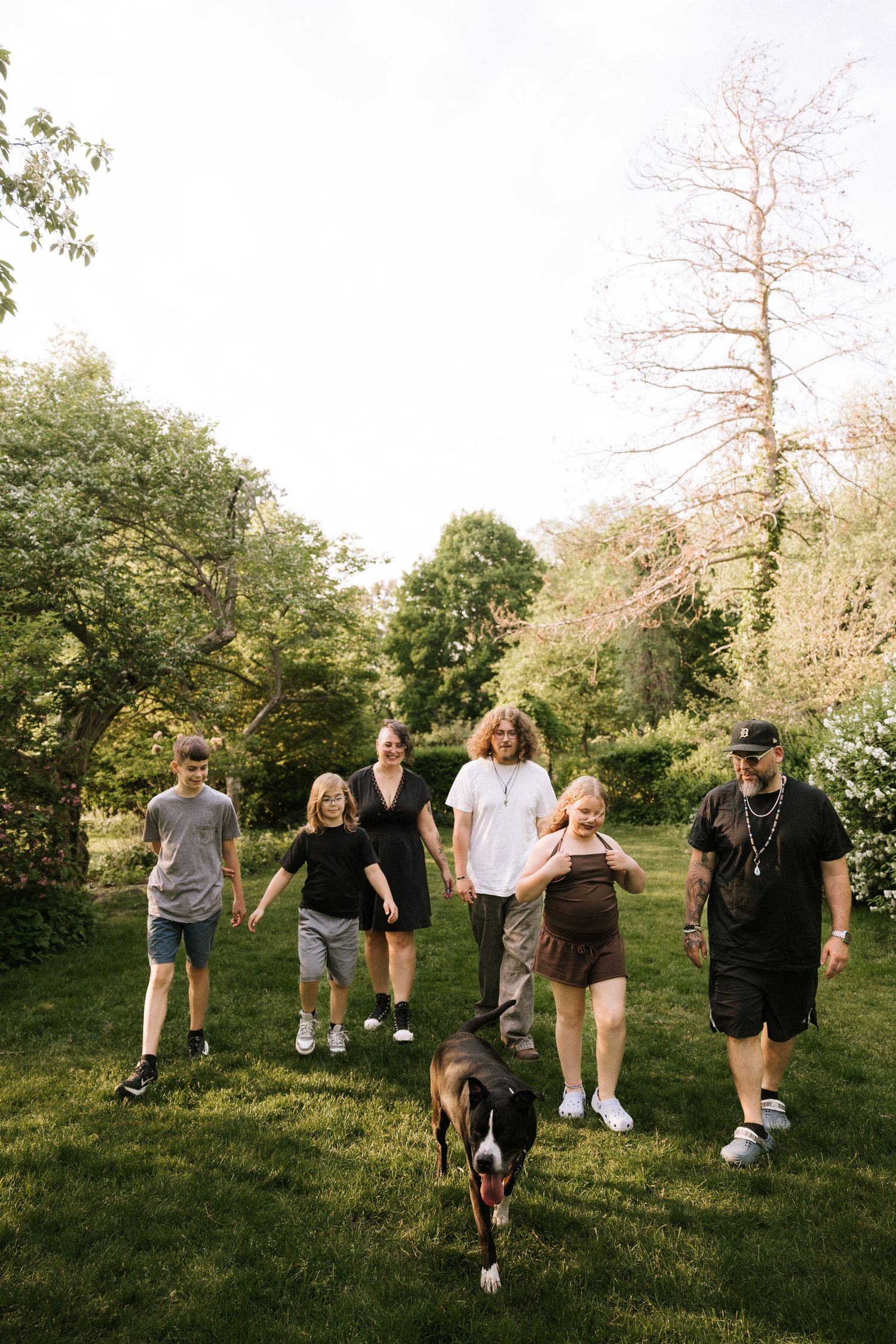 Family with dog walking together on lush green lawn surrounded by trees on sunny day.