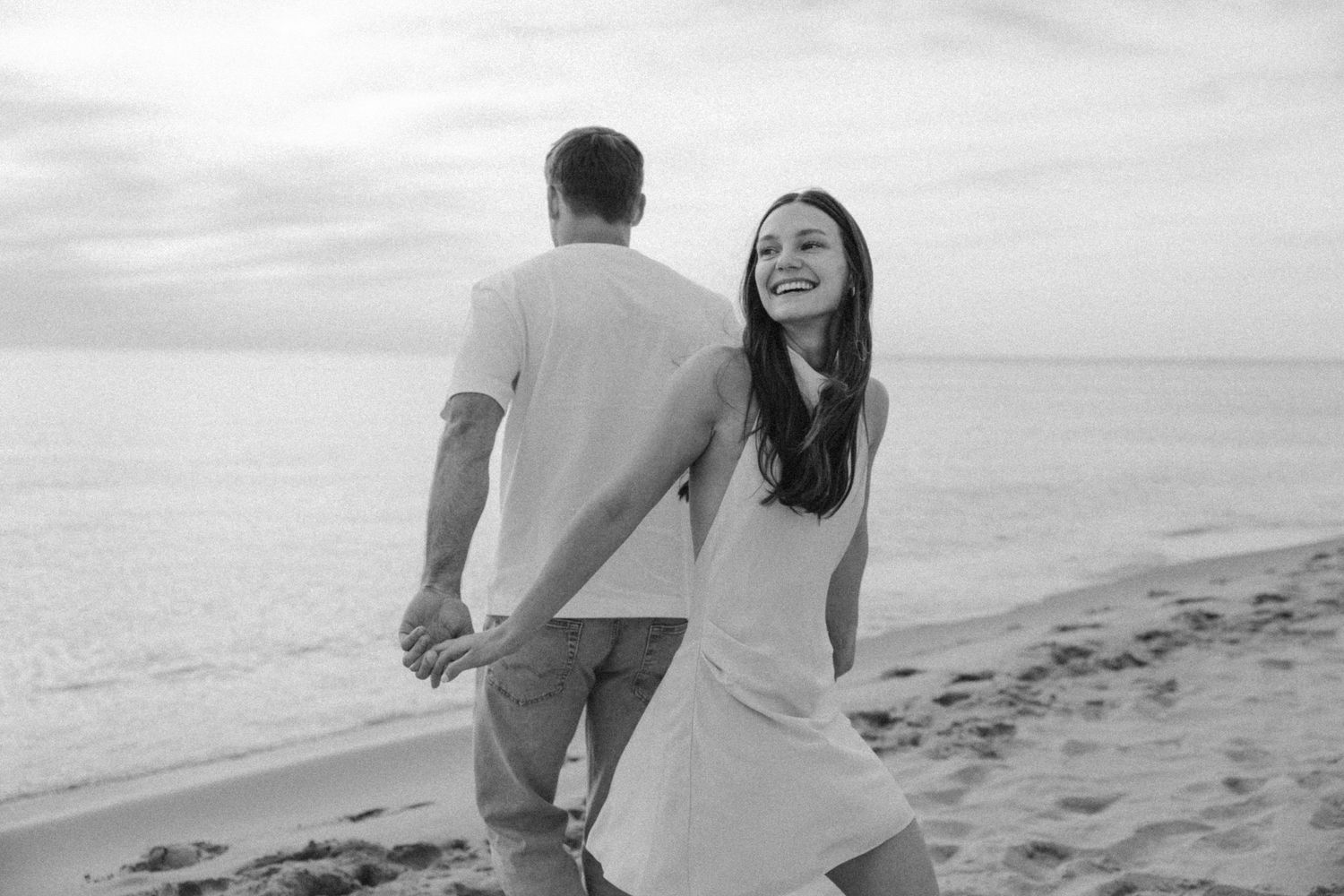 Black and white photo of a joyful couple holding hands and walking along a beach at sunset.