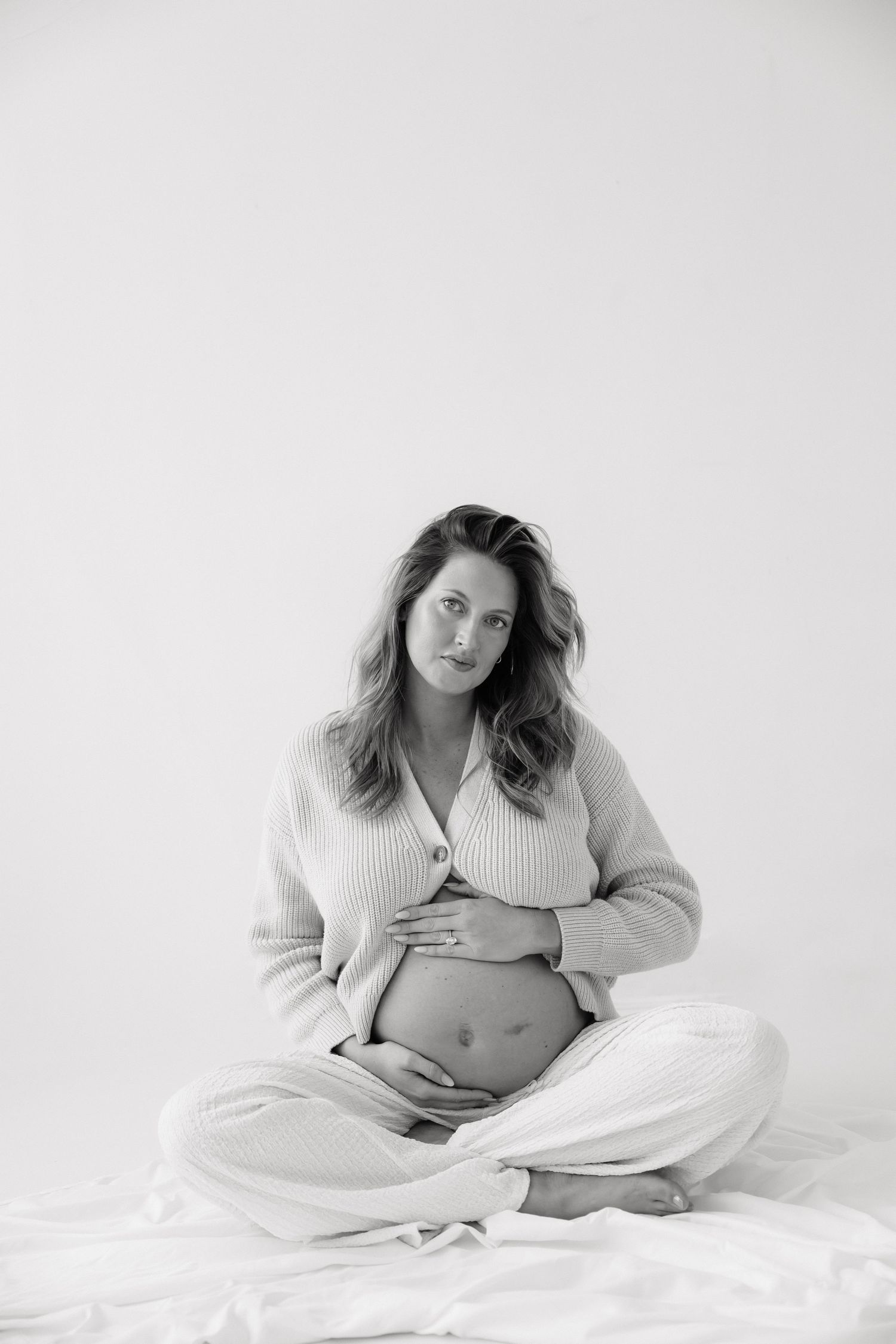 Black and white photo of a pregnant woman in a knit cardigan sitting cross-legged, cradling her baby bump while looking at camera.