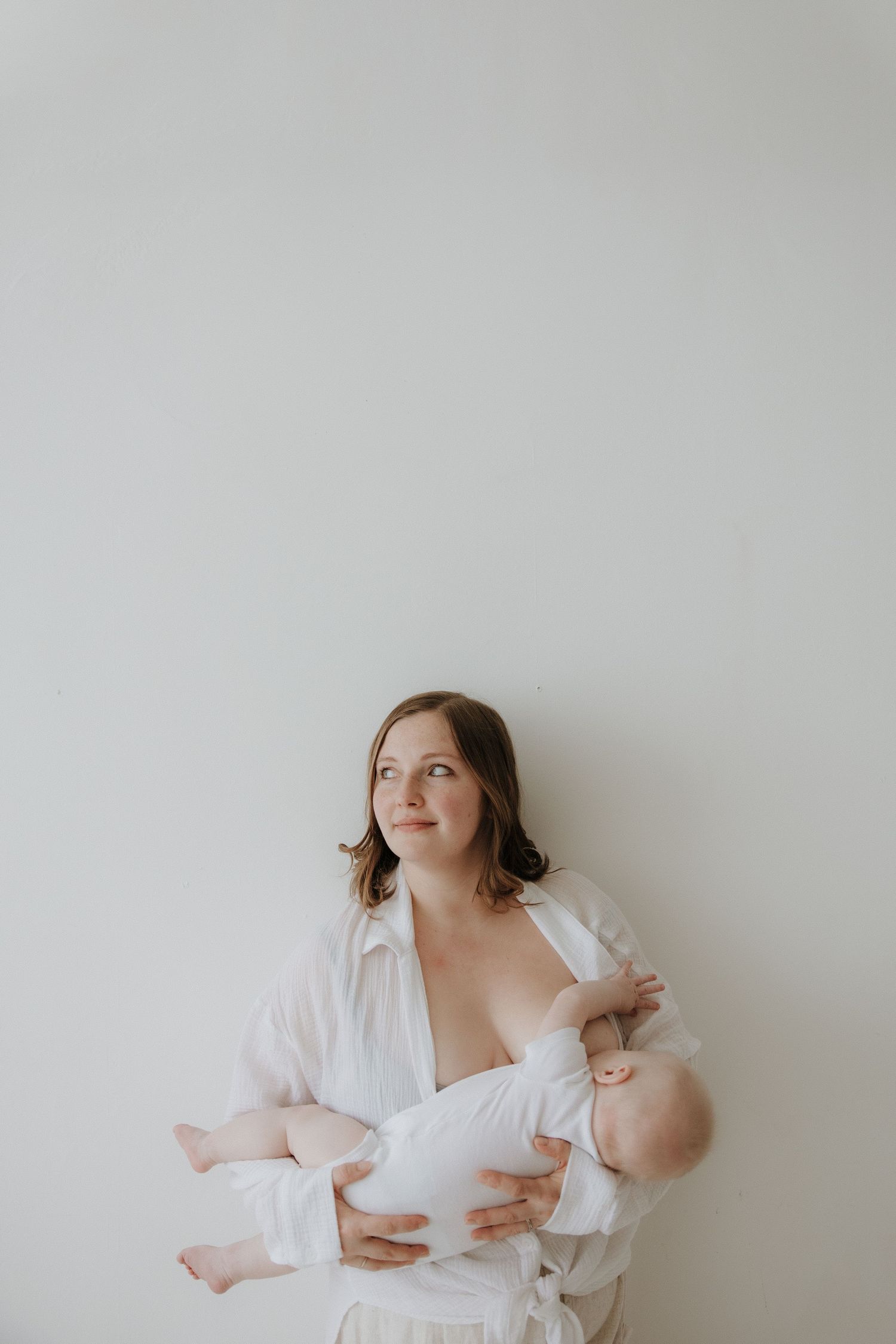 A mother in a white shirt breastfeeds her newborn baby while gazing upward against a soft gray background.