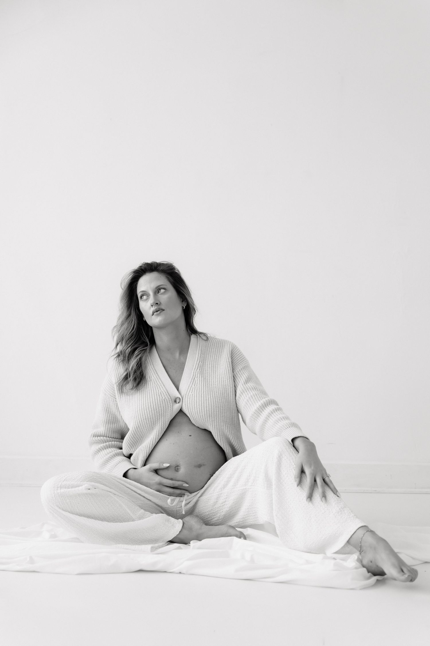 Black and white maternity photo of a pregnant woman sitting on the floor in a knit cardigan, gazing upward thoughtfully.