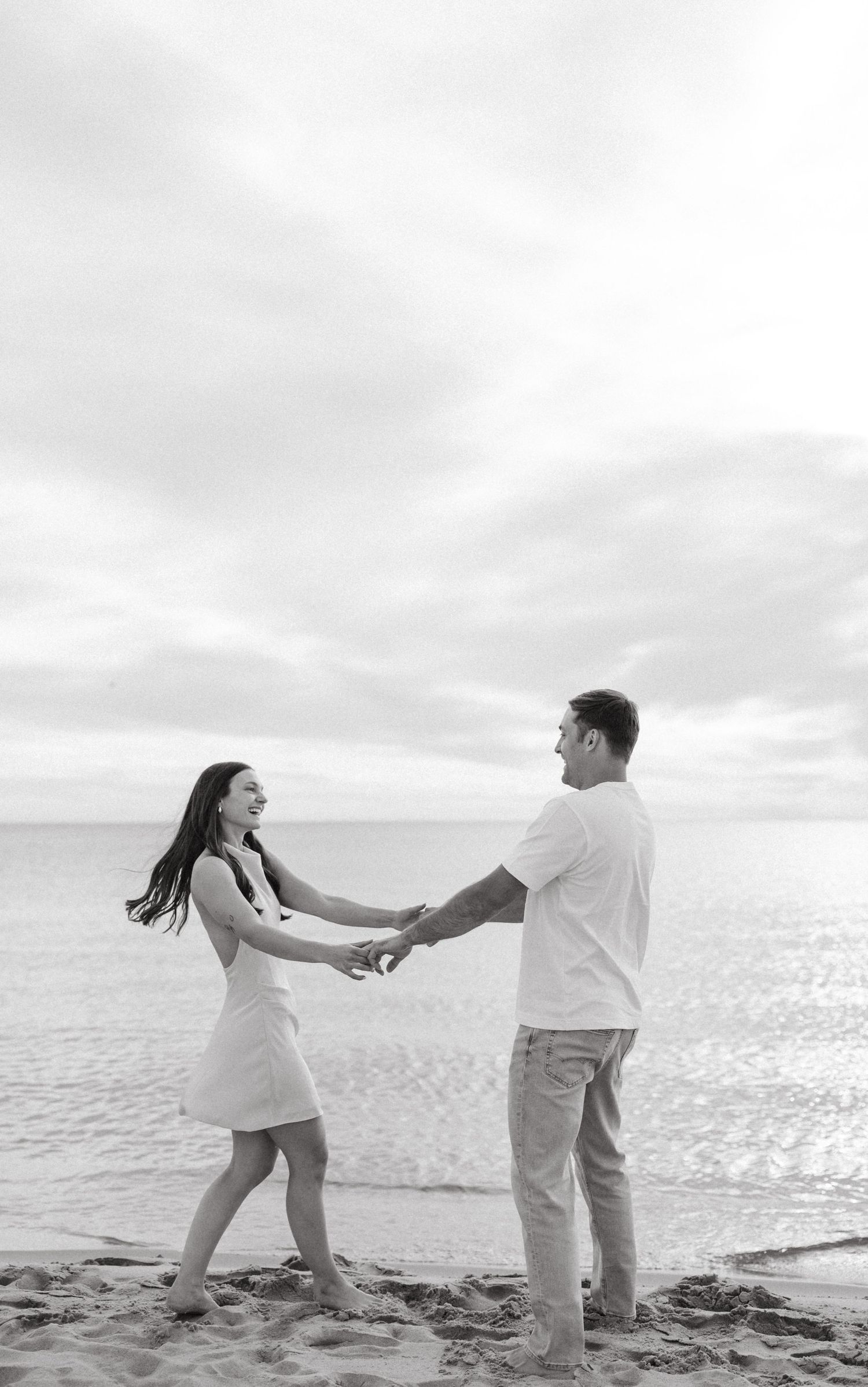 Black and white photo of couple holding hands and dancing playfully on sandy beach by the ocean.