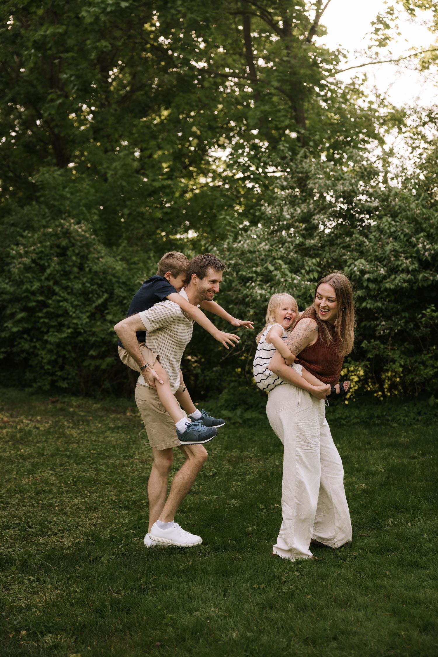 A joyful family plays in a lush green park, dad giving piggyback ride to son while mom holds toddler.