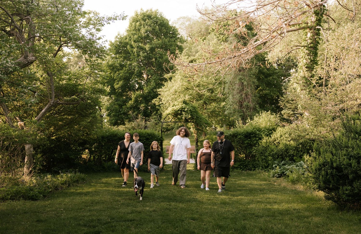 Group of friends walking together across a lush green garden surrounded by tall trees on a sunny day.