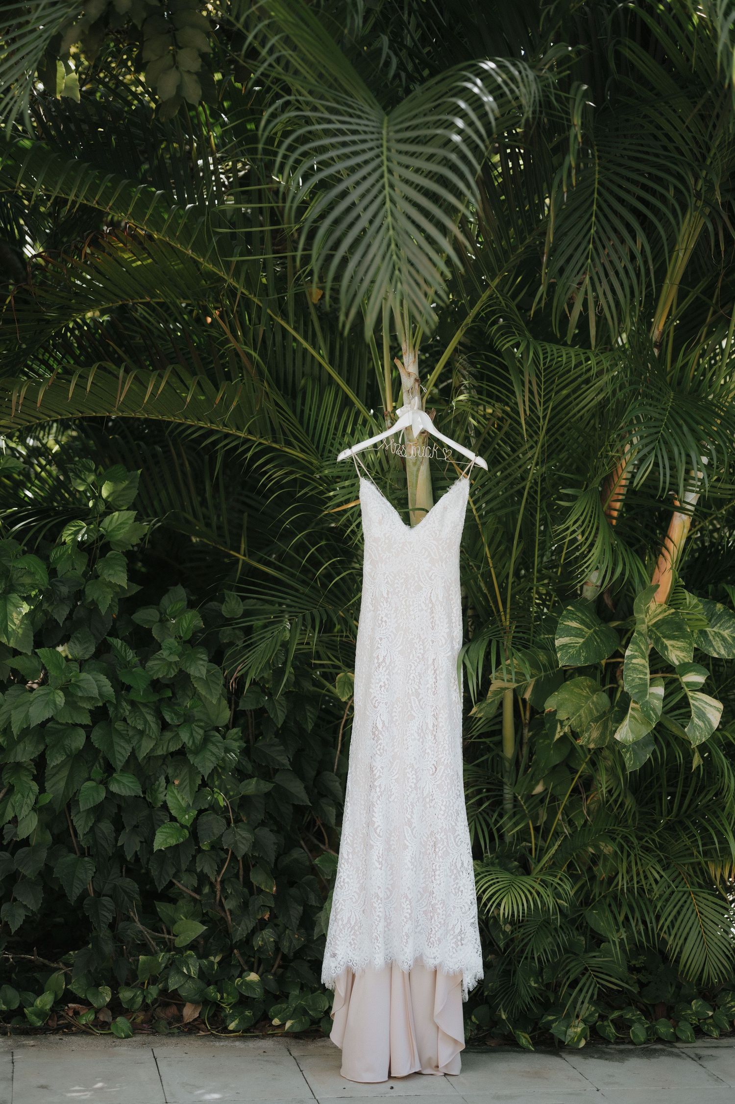 White lace wedding dress displayed against lush tropical palm fronds and greenery.