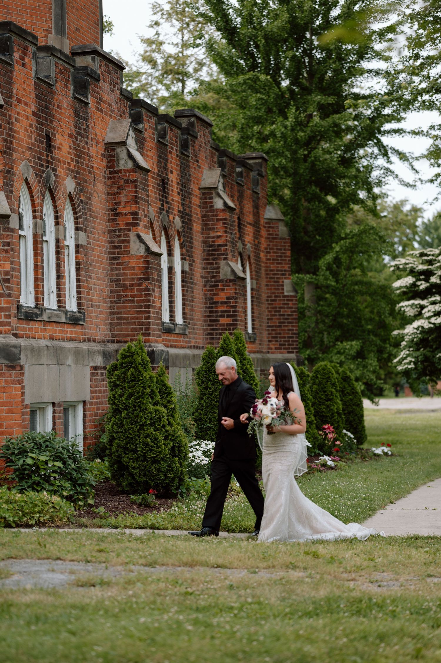 Romantic first look moment outside historic brick building with Gothic windows and lush greenery.