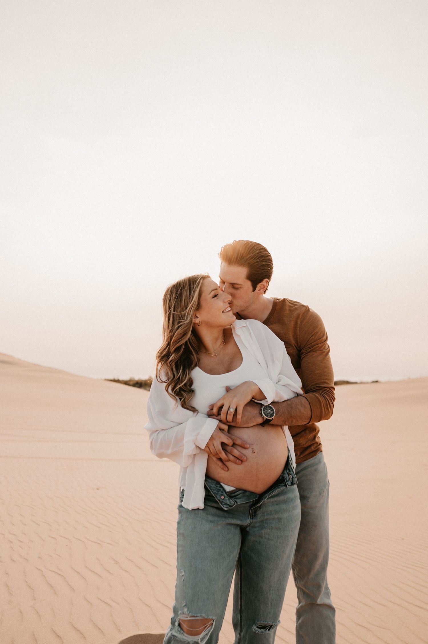 Expecting couple embraces in desert setting during sunset maternity photoshoot.