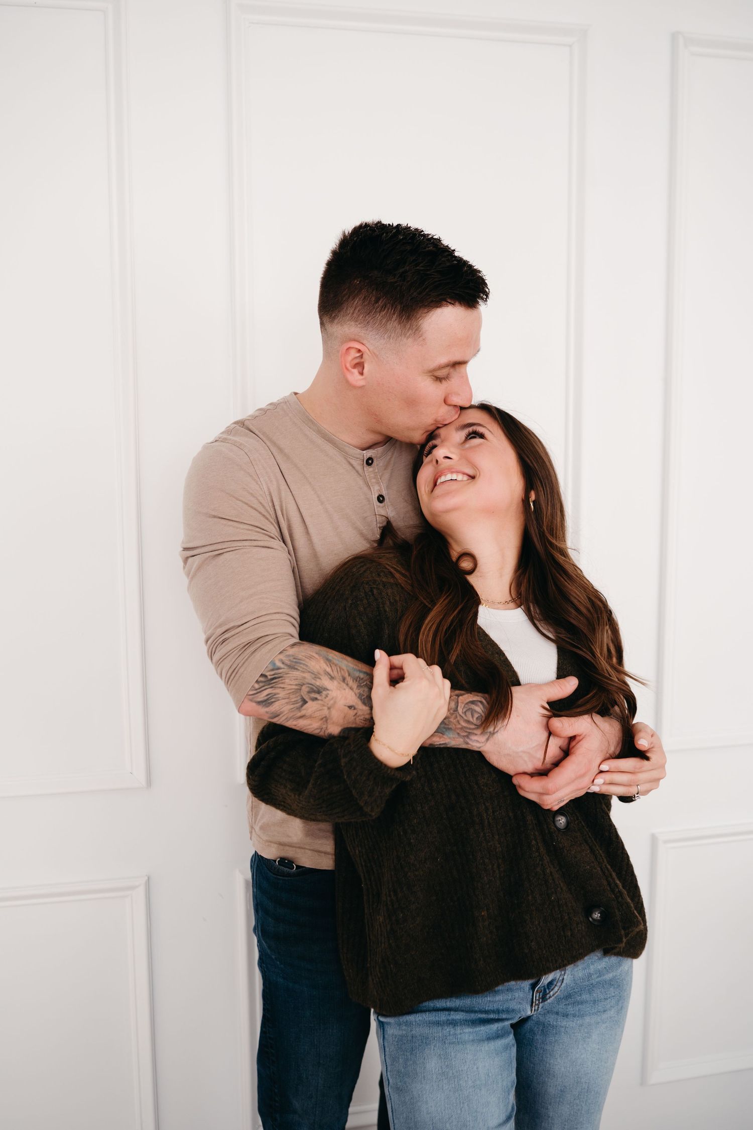 A couple shares a tender moment embracing and laughing together against a white paneled wall.