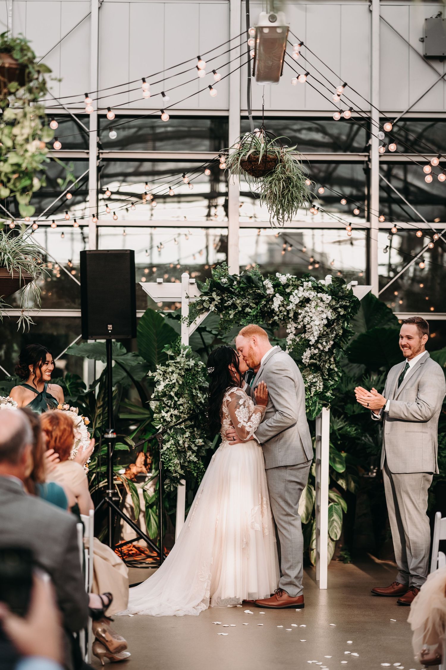 First kiss as newlyweds under string lights in greenhouse wedding venue.