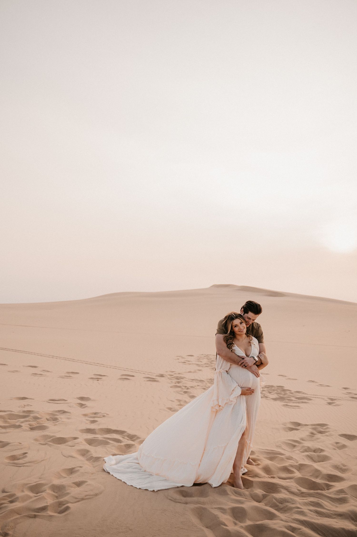 A romantic couple embraces on sand dunes during sunset, with the bride wearing a flowing white dress.
