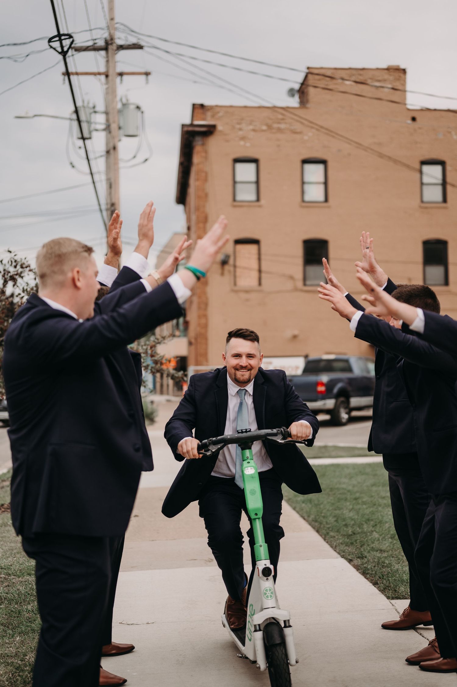 Wedding party member rides electric scooter while others cheer on sidewalk.