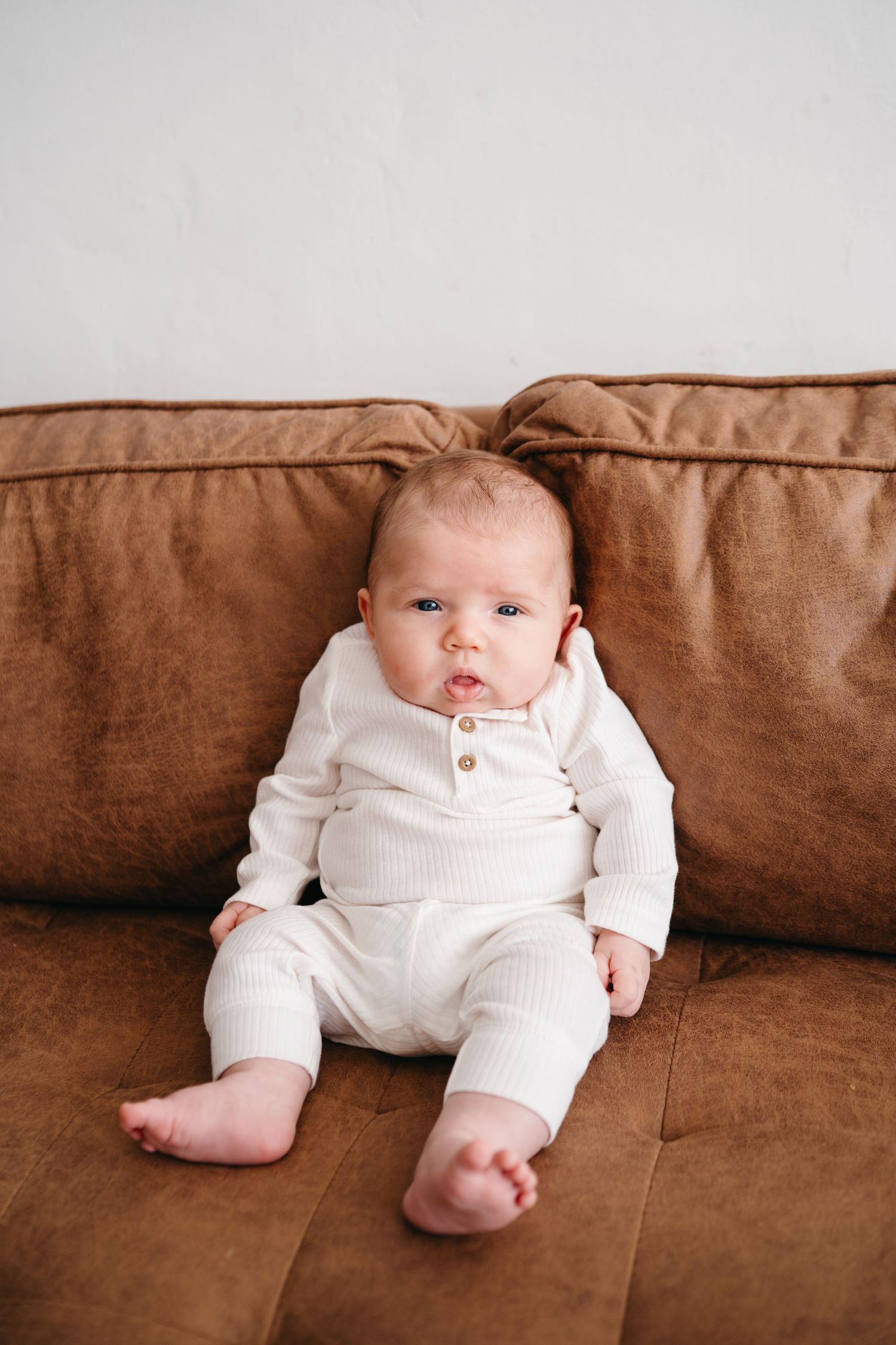 Baby wearing white knit outfit sits on brown leather couch with bare feet showing.