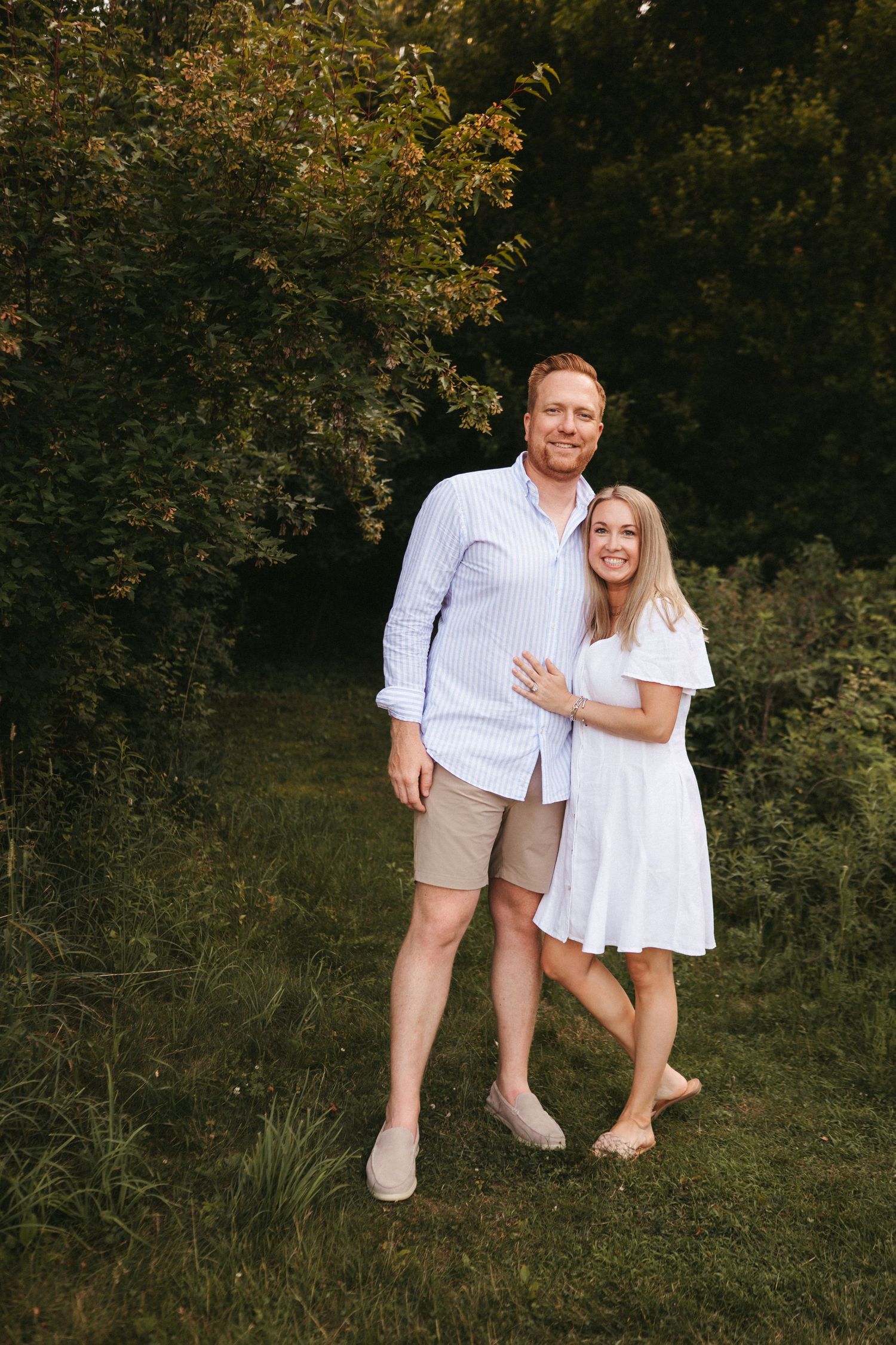 Couple in coordinated white outfits pose together on grassy lawn during golden hour with trees in background.