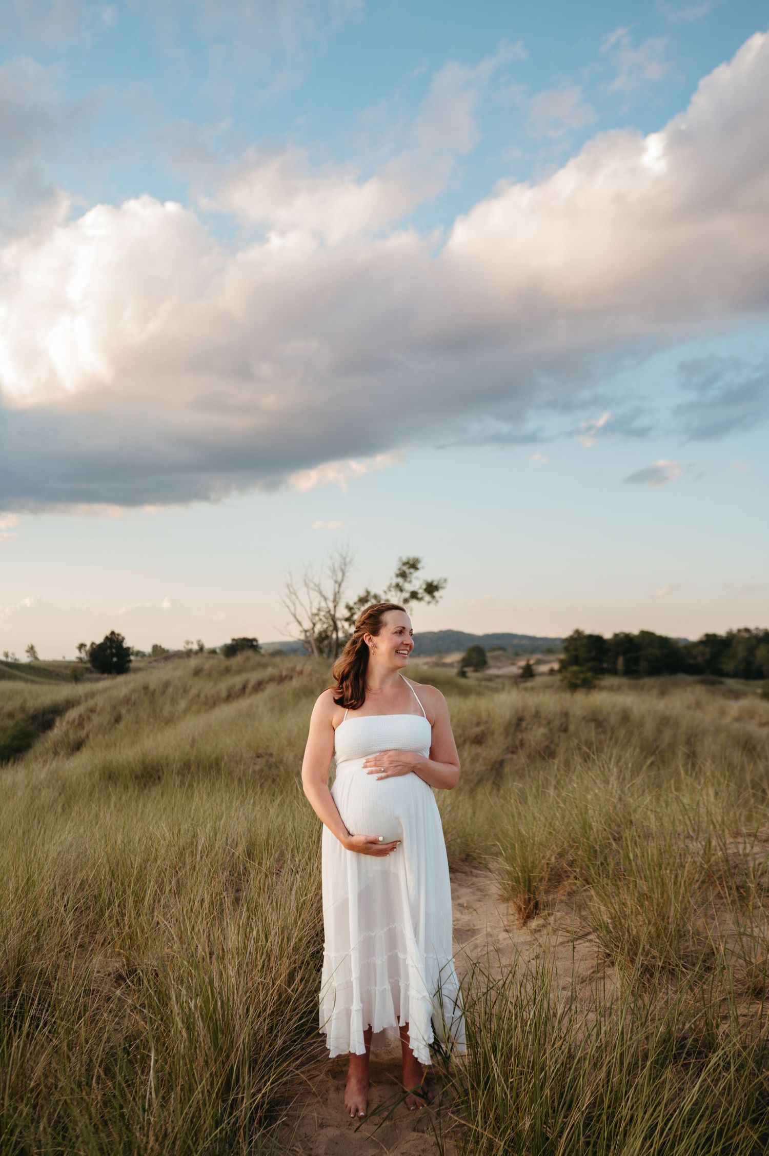 A figure in a flowing white dress stands on a grassy hilltop path at sunset with mountains in the distance.