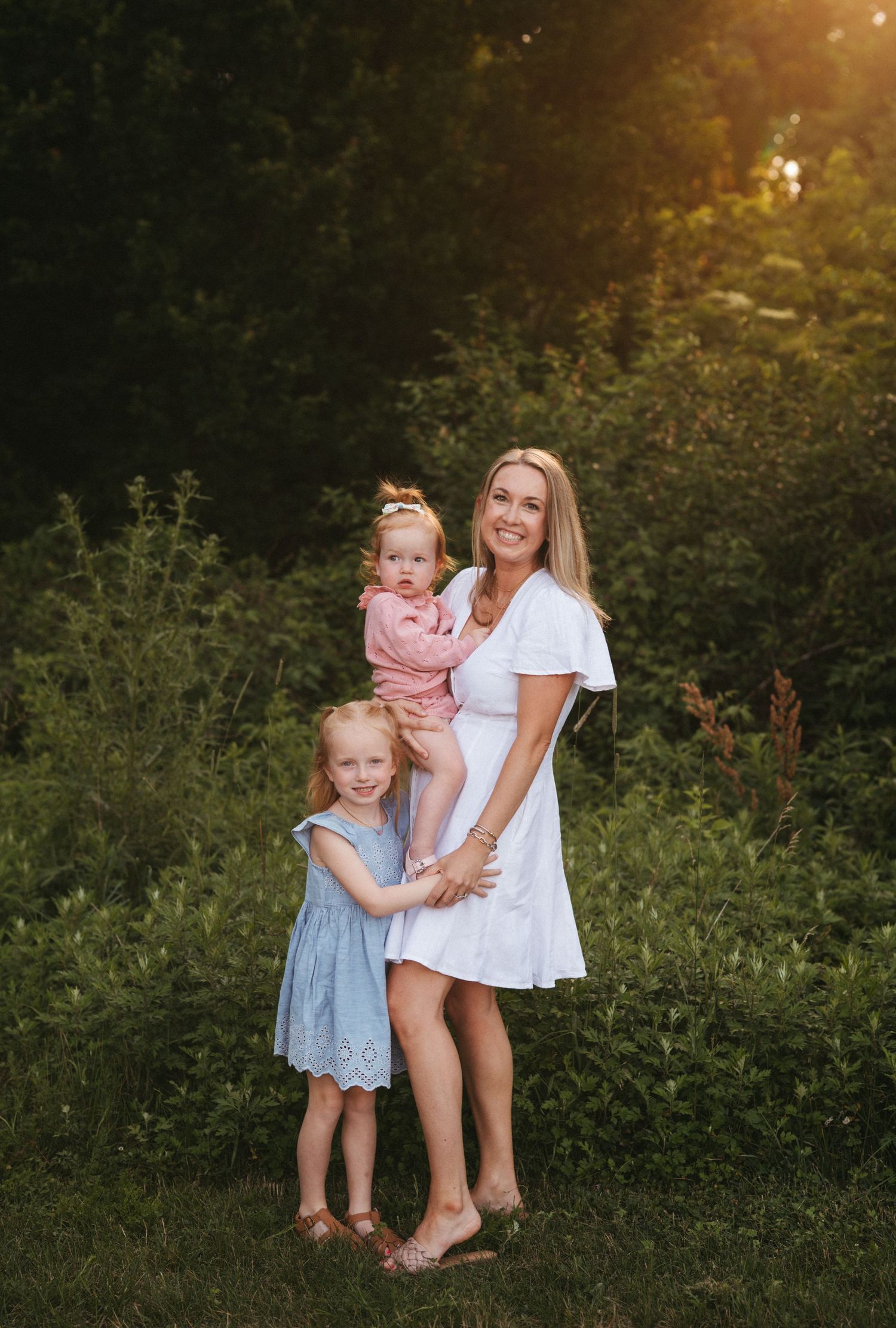 Three children in coordinating outfits pose together during a sunset photo session in a garden.