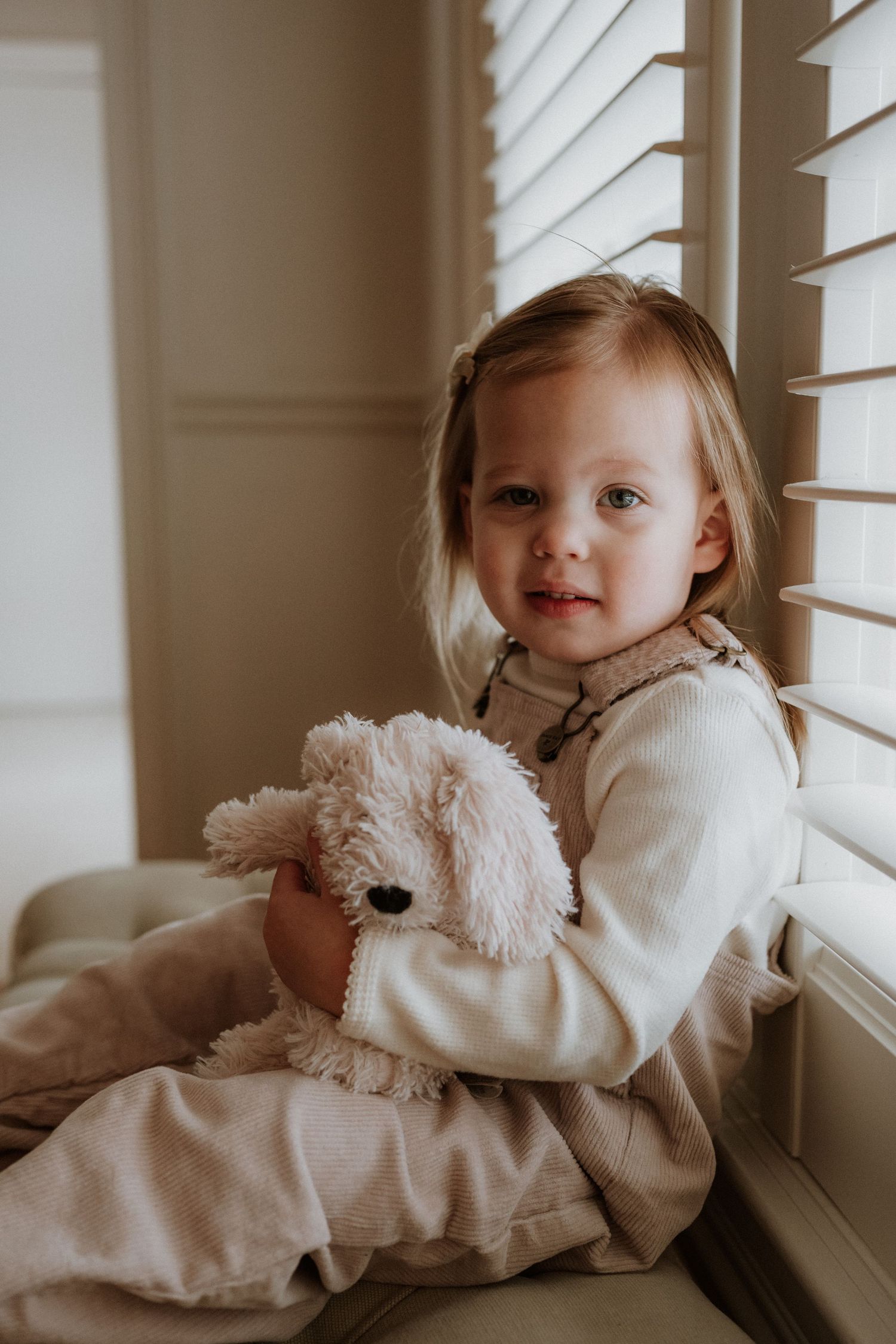 A toddler in beige clothing sits by a window holding a plush stuffed dog toy.