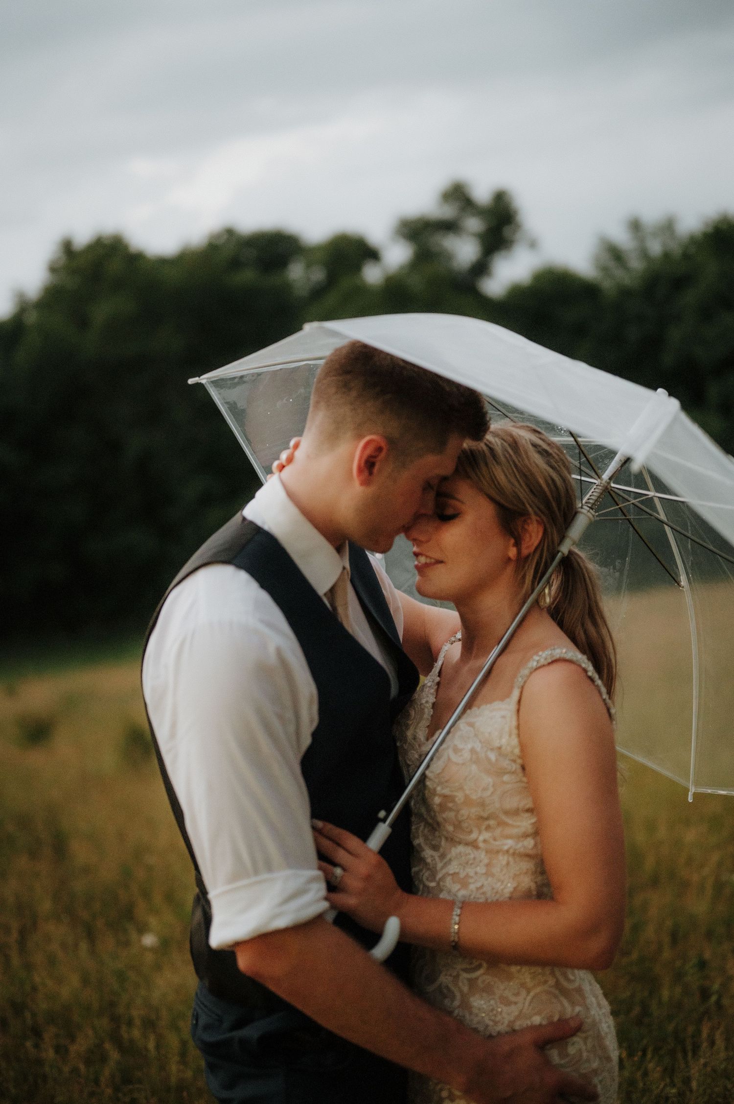 Couple shares intimate moment under clear umbrella in rustic field setting.
