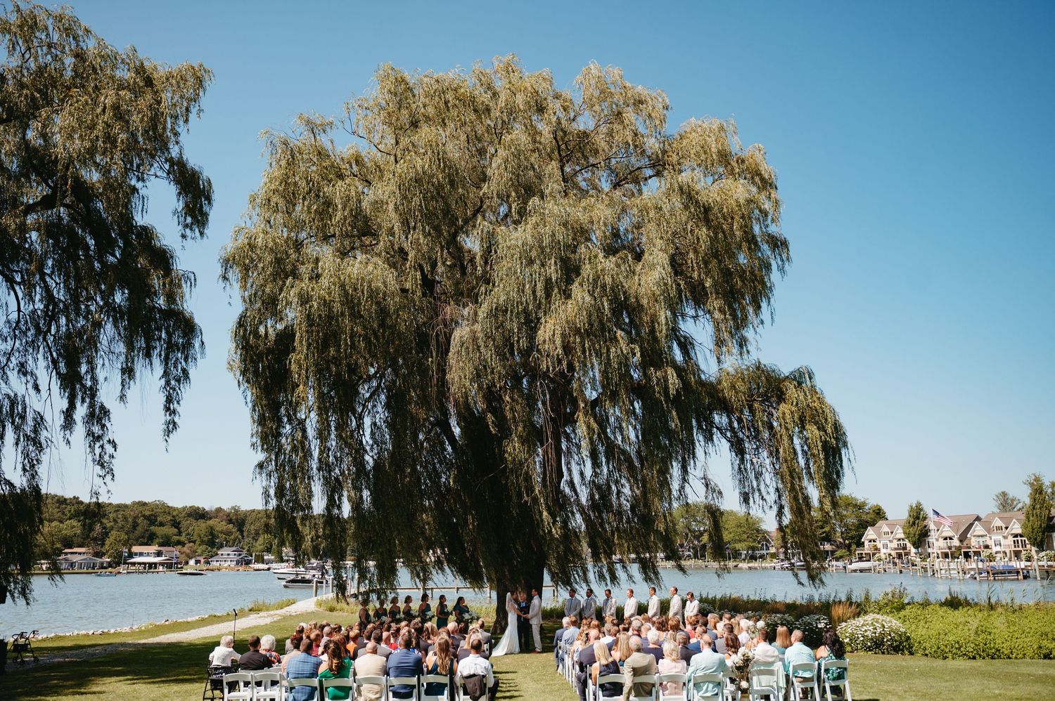 Panoramic view of a waterfront wedding ceremony under weeping willow trees.