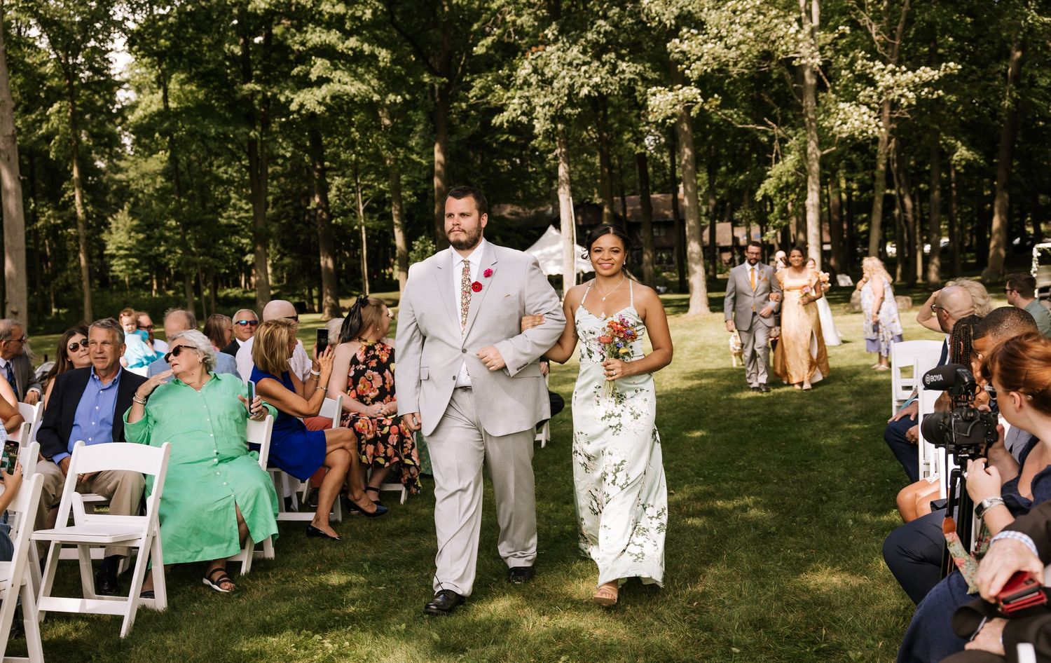 Wedding guests gather on white chairs under tall trees during an outdoor ceremony.