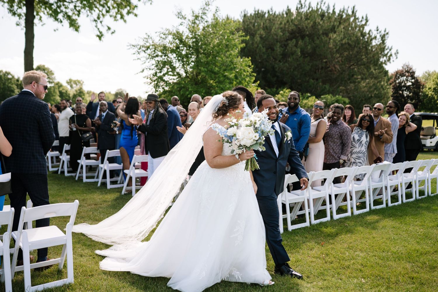 Wedding guests seated in white chairs on a manicured lawn during an outdoor ceremony.