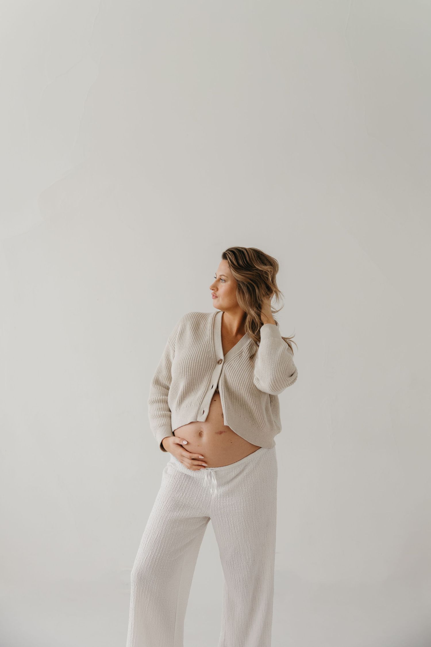 Expectant mother in cream knit cardigan and white ribbed pants gently holding baby bump in studio setting.
