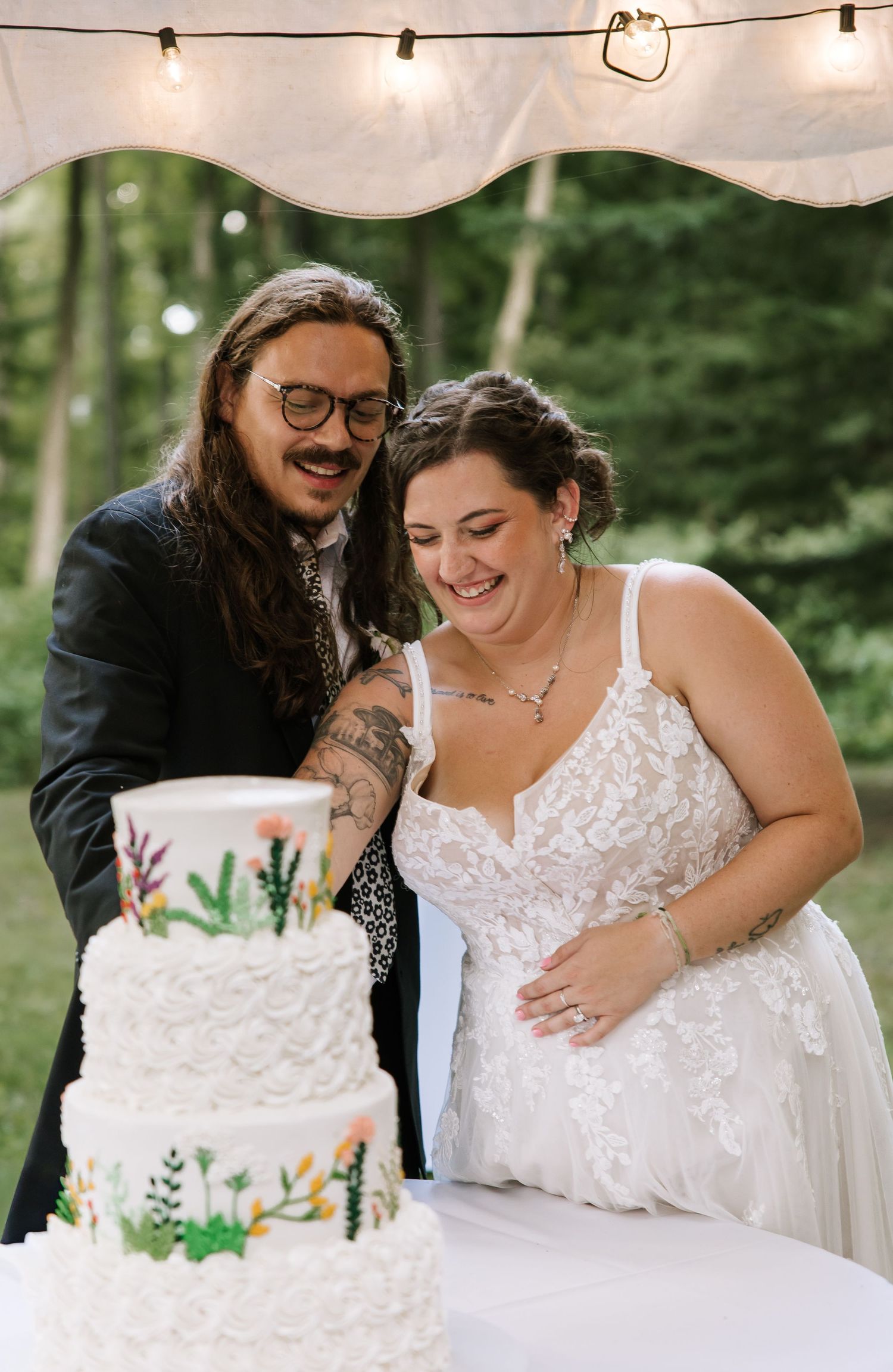 Happy couple cutting their white tiered wedding cake together under string lights.