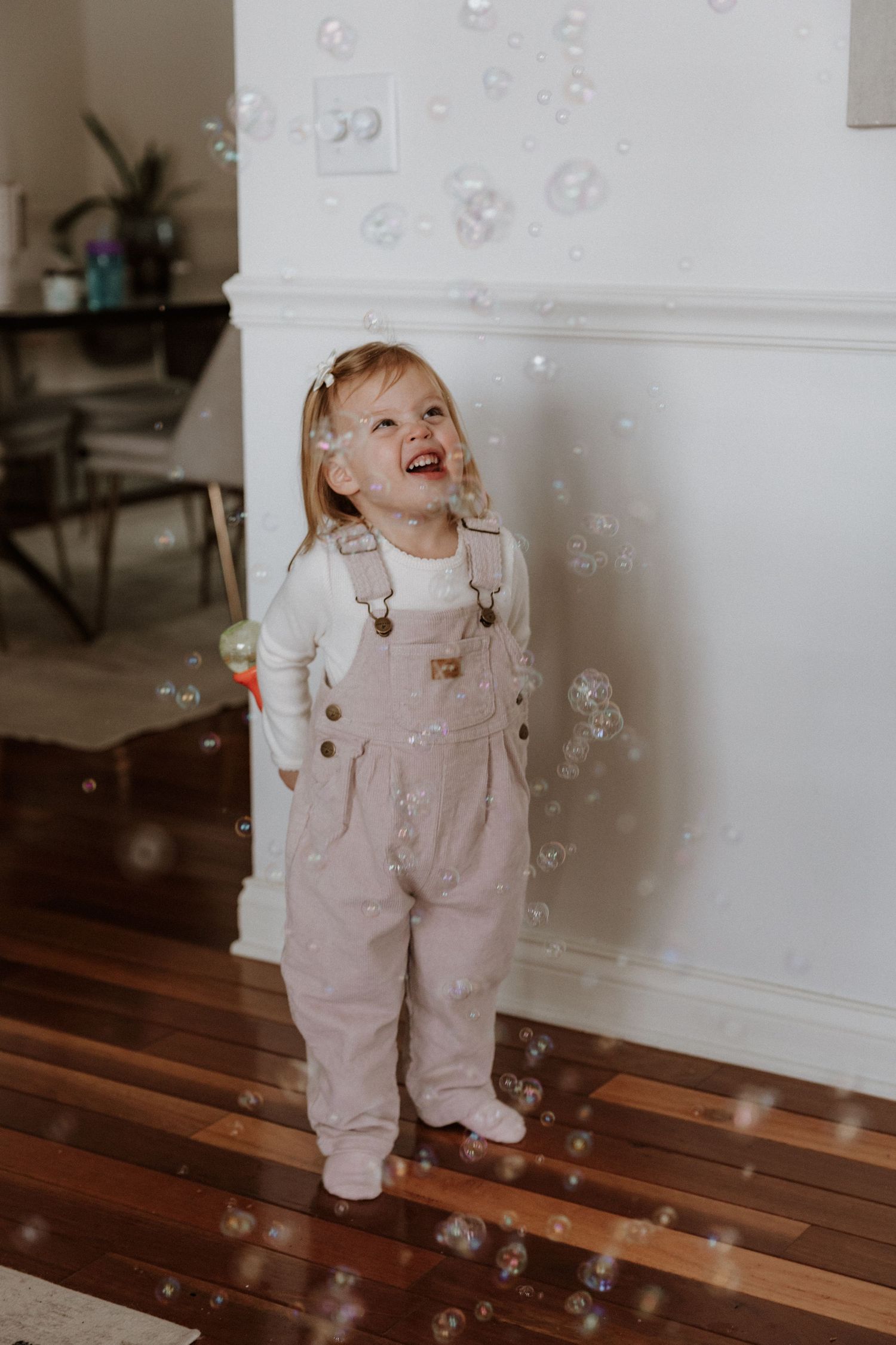 Toddler in overalls laughing while playing with bubbles indoors.
