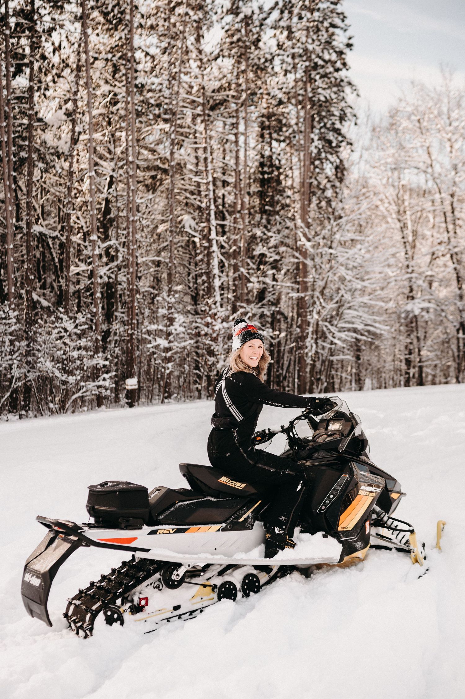 Person riding a black and orange snowmobile through a snow-covered forest.
