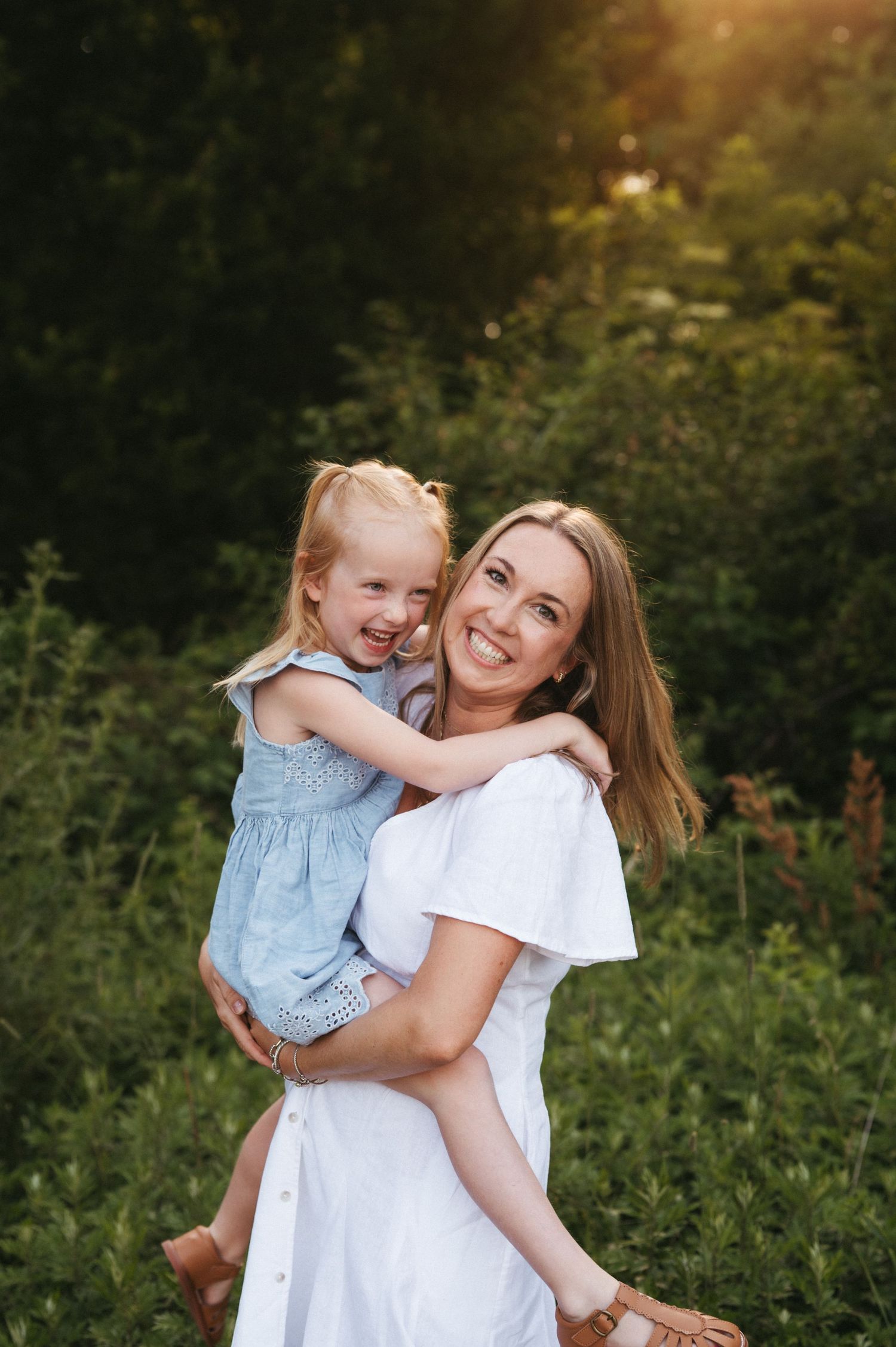 Joyful outdoor portrait of parent and child embracing in white outfits against greenery.