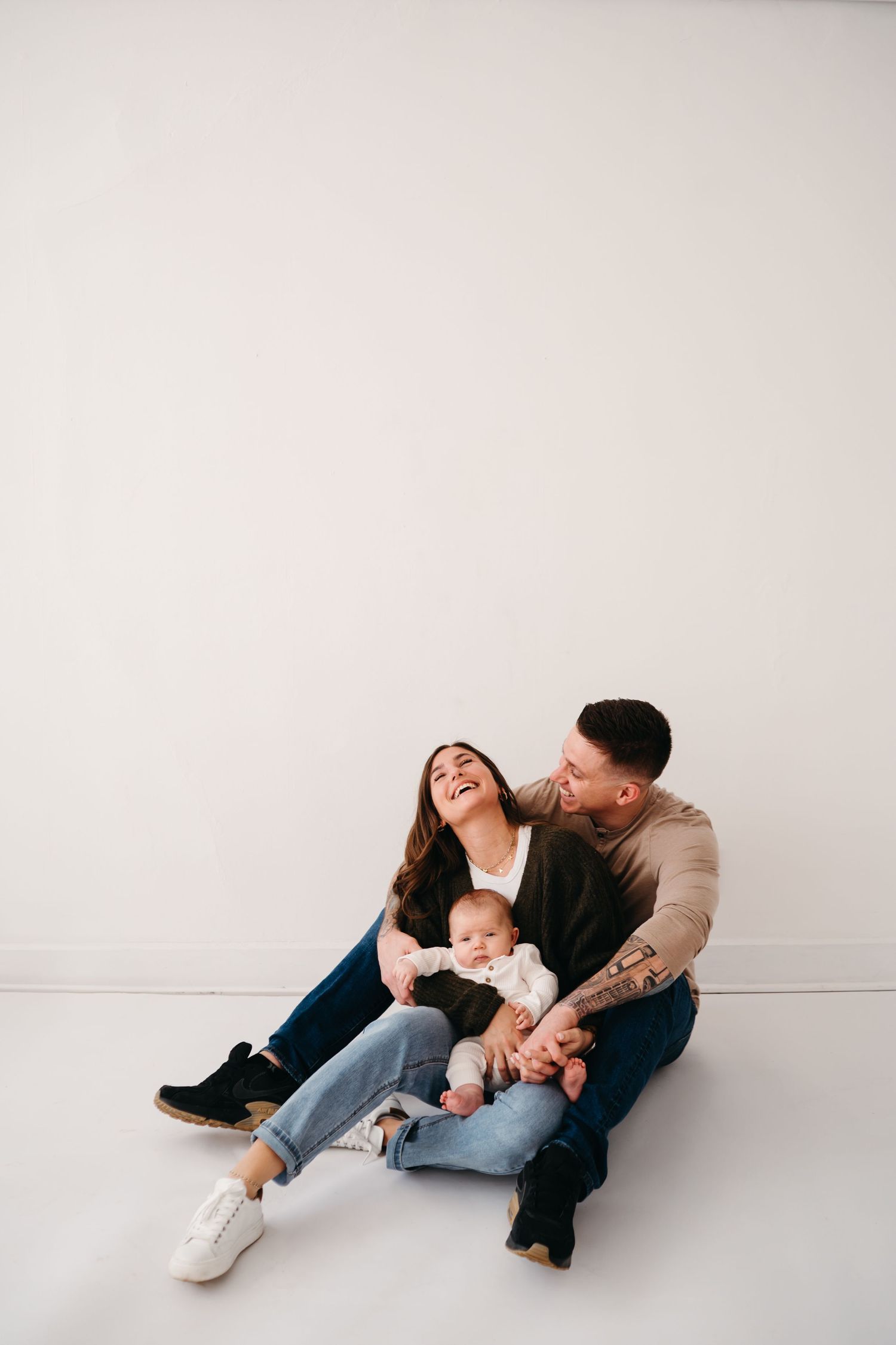 A couple and their baby share a joyful moment while sitting on white studio floor.