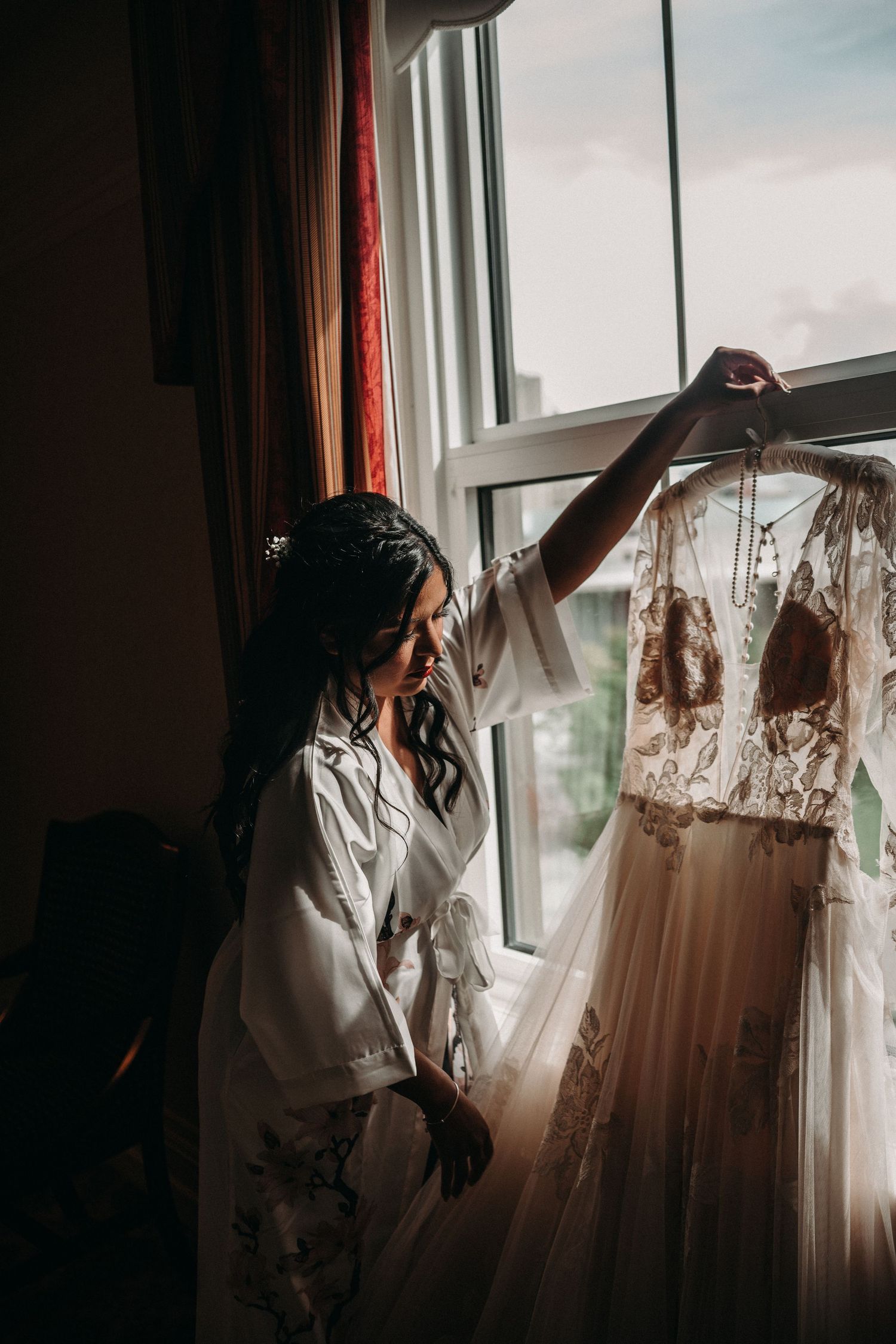 Silhouetted figure admires wedding dress hanging by window in morning light.
