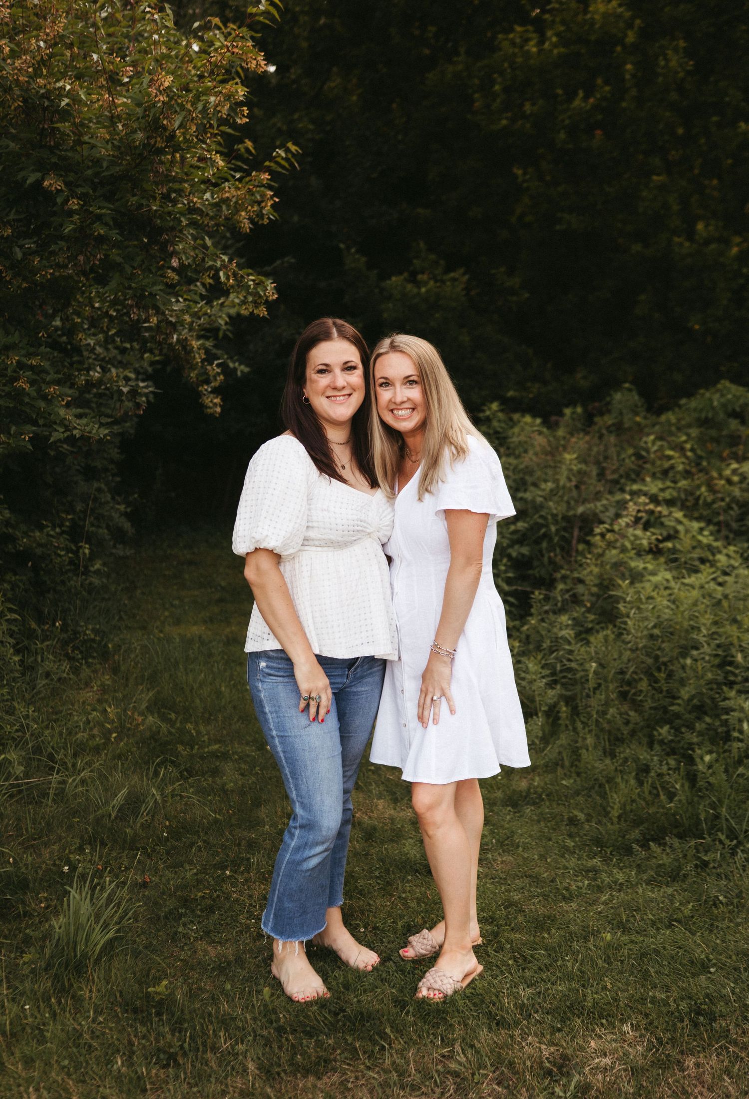 Two people wearing white tops pose together outdoors in front of green foliage on a summer day.