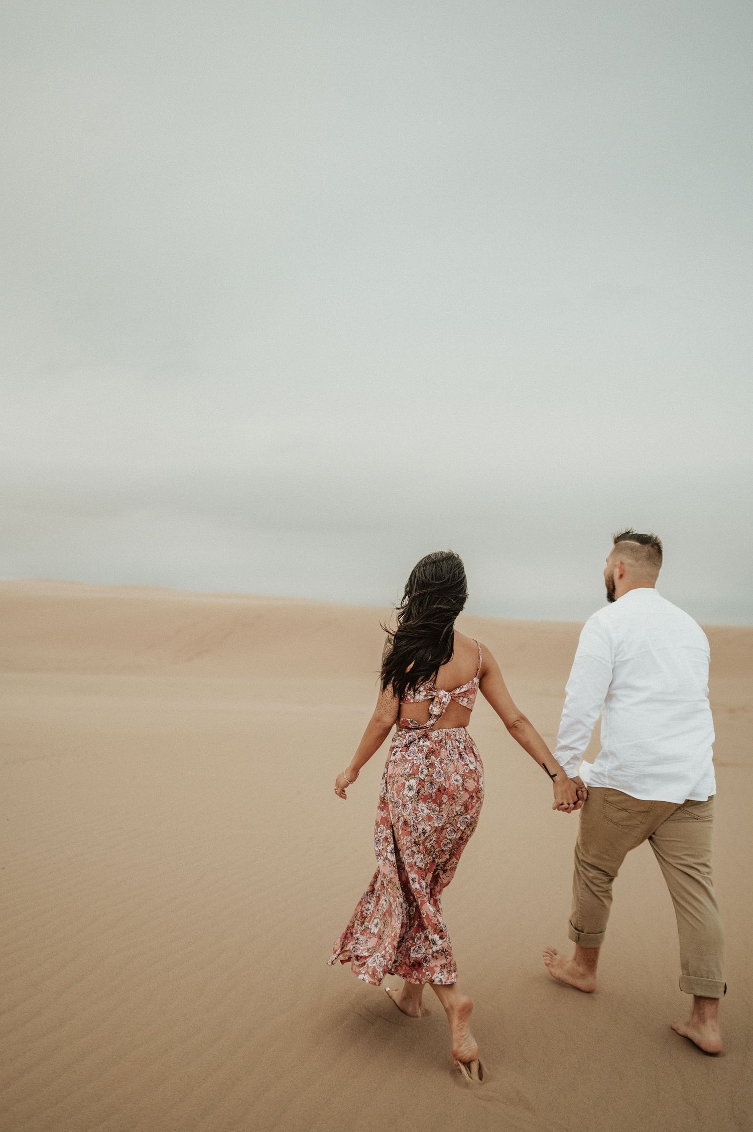 A couple walks barefoot hand-in-hand across desert sand dunes under an overcast sky.