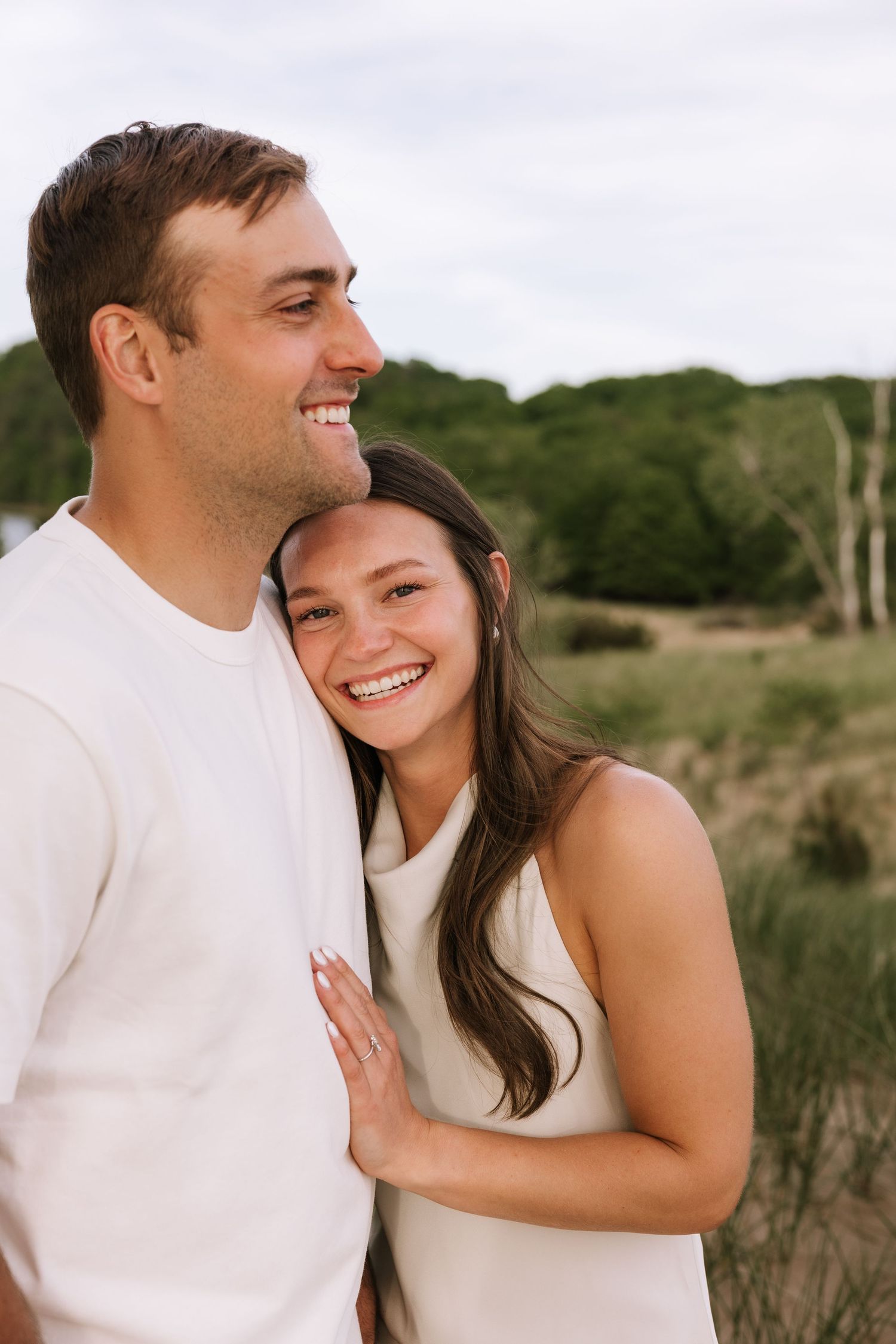 Smiling couple standing together outdoors, woman in white dress leaning against mans chest.