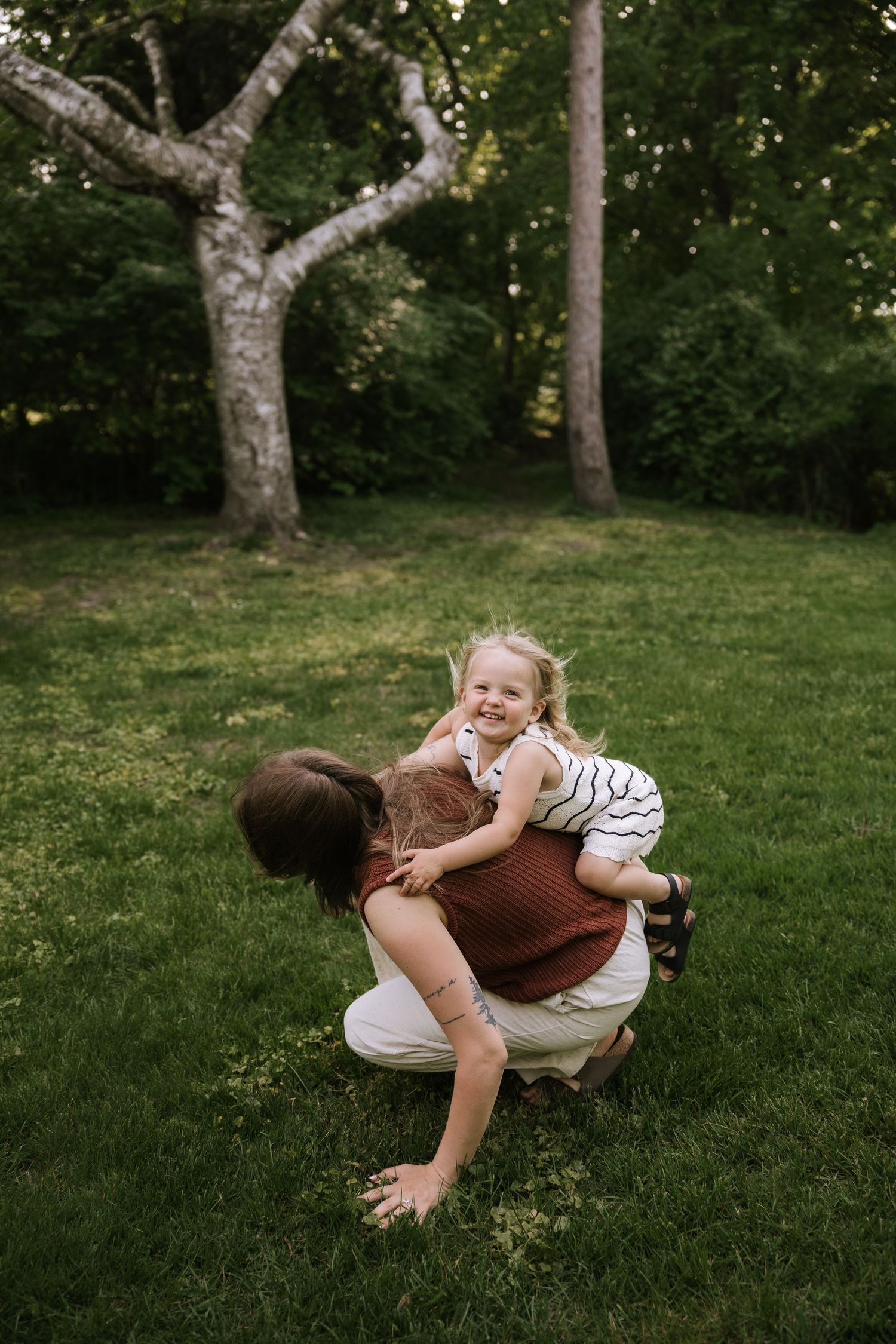 A mother crouches on green grass hugging her laughing toddler daughter outdoors in a lush park setting.