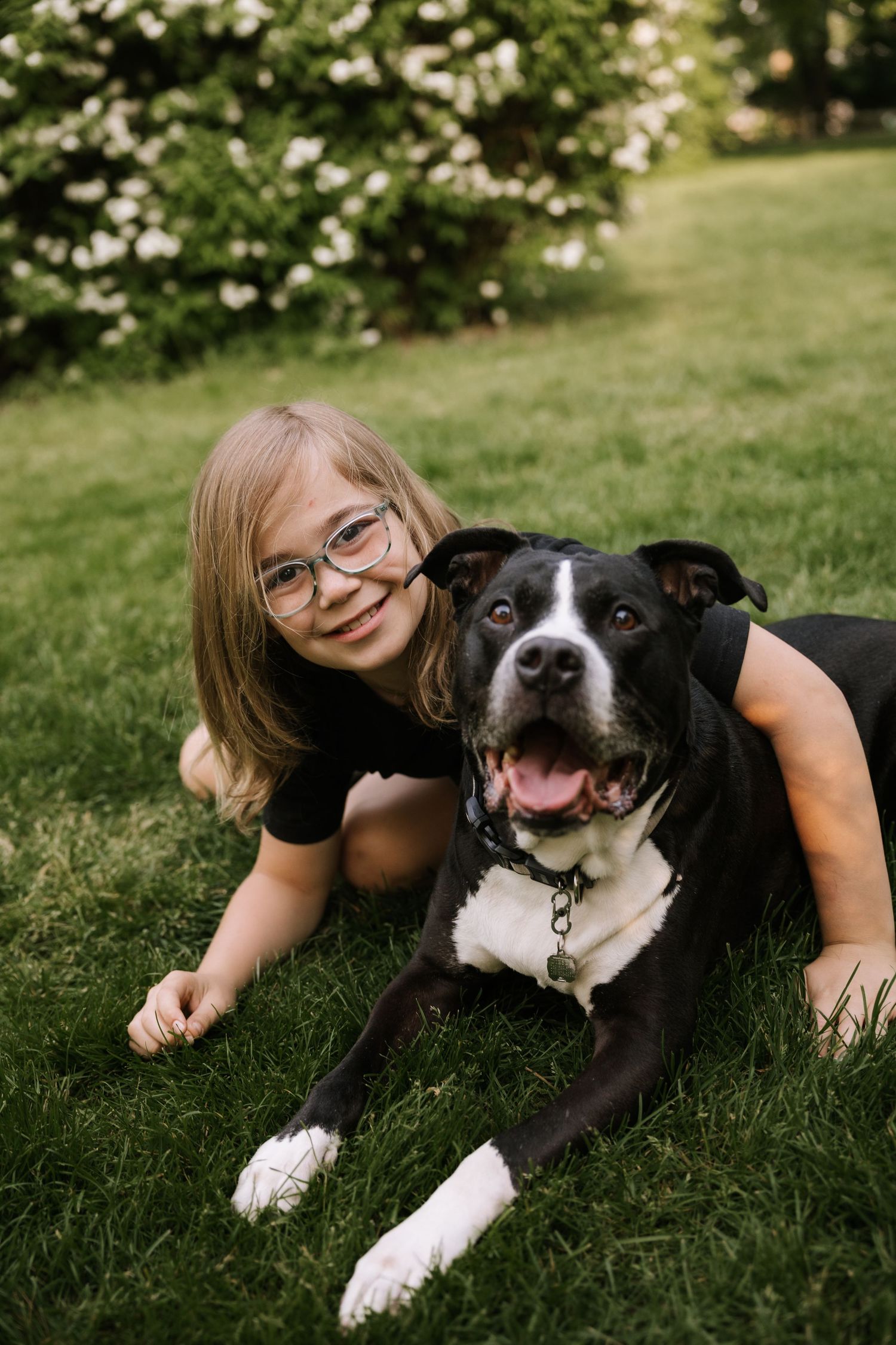 Young girl with glasses smiles while lying on grass with a black and white dog outdoors.