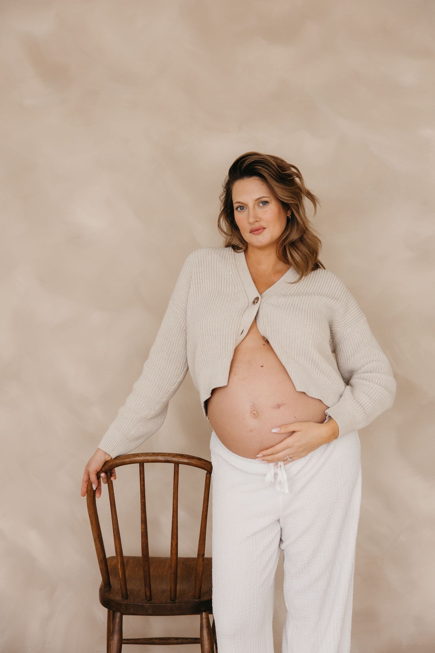 Pregnant woman cradling baby bump in open beige cardigan and white pants beside wooden chair.
