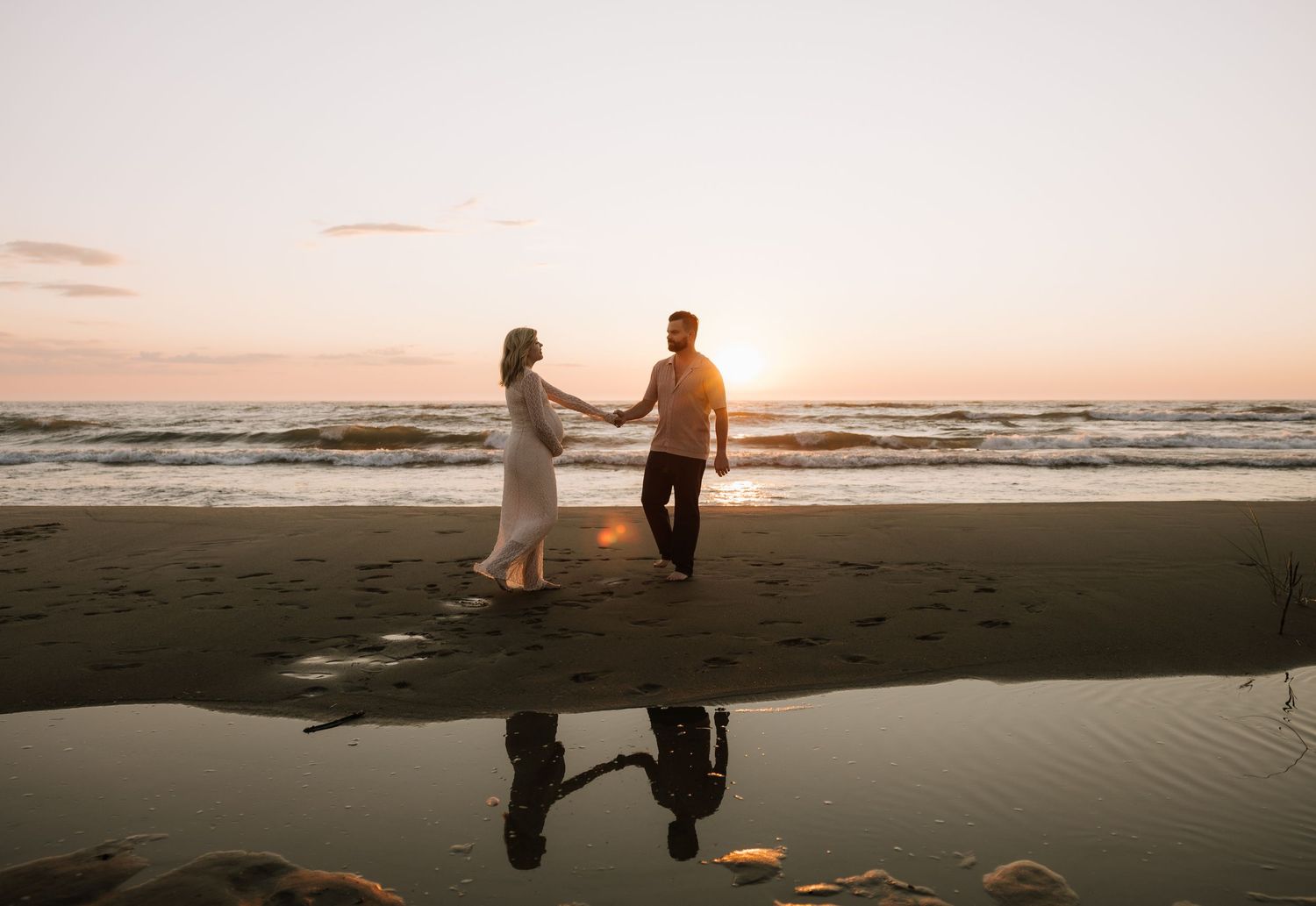 Couple holding hands on a beach at sunset with their reflection visible in the wet sand by the ocean waves.