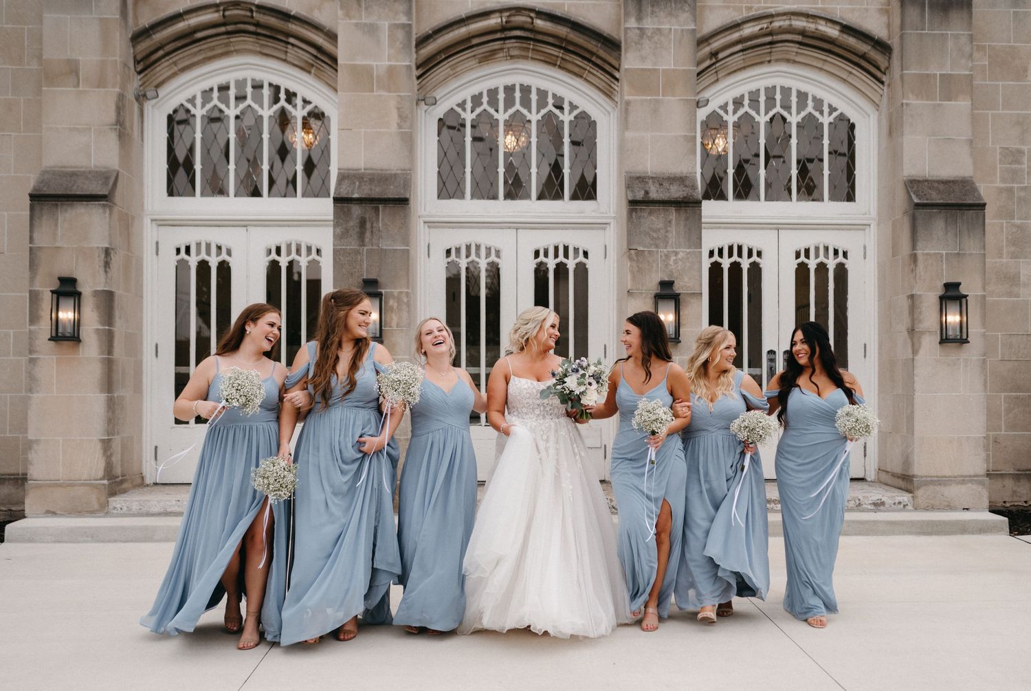 Wedding party in dusty blue bridesmaid dresses poses playfully against gothic stone church architecture.