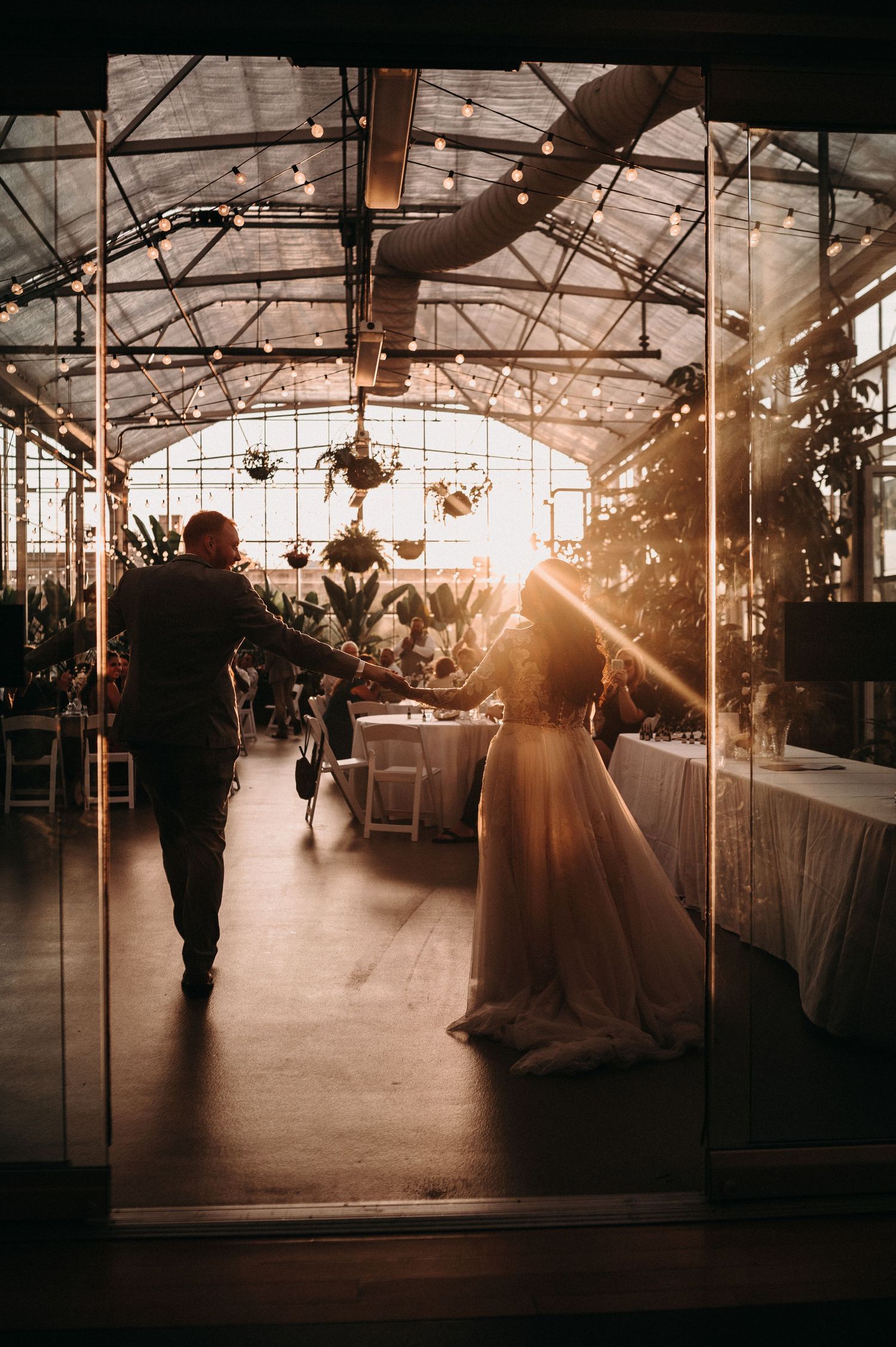 Wedding couple in silhouette walking through sunlit greenhouse with industrial architecture.