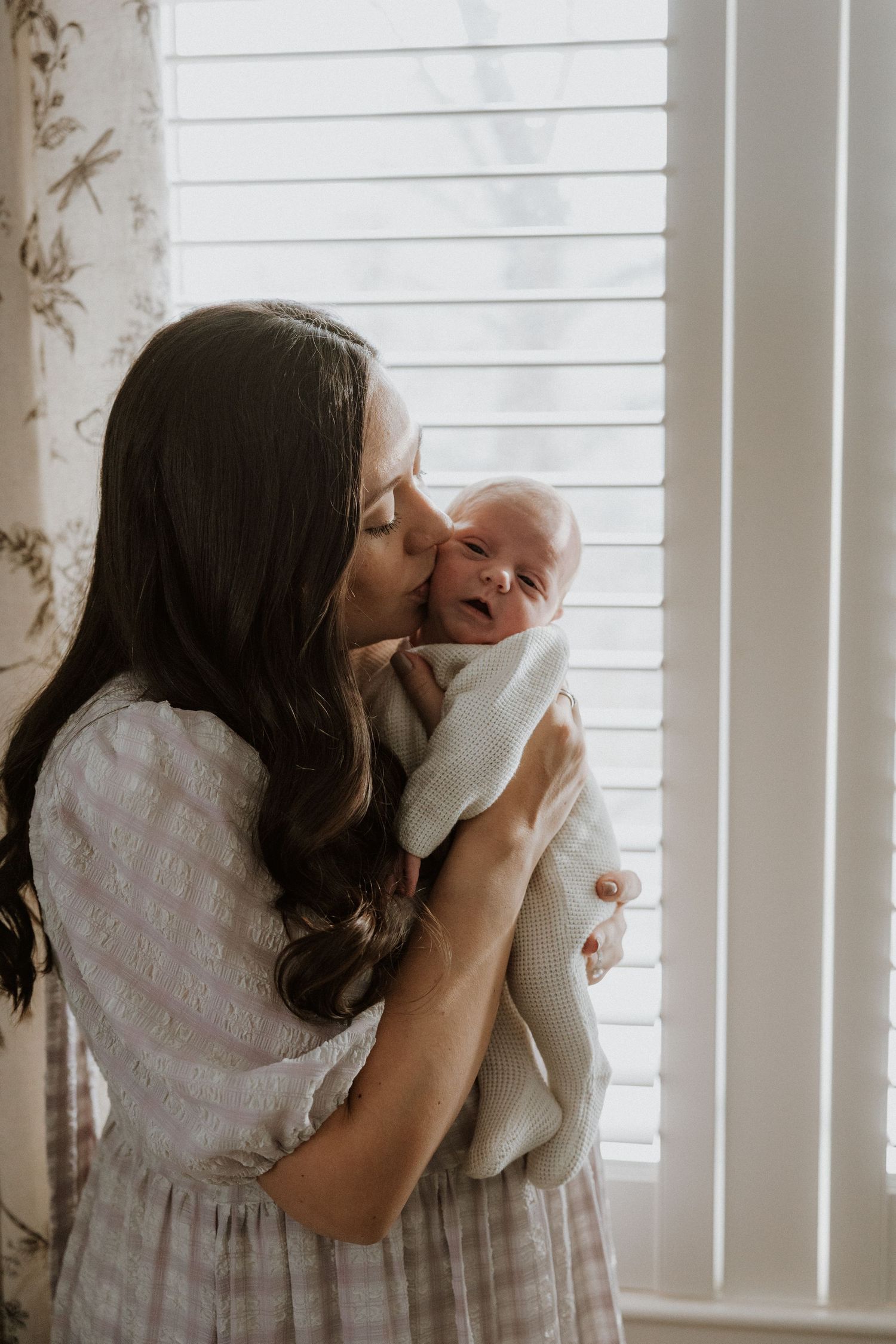 Tender moment between parent and baby by sunlit window with white blinds.