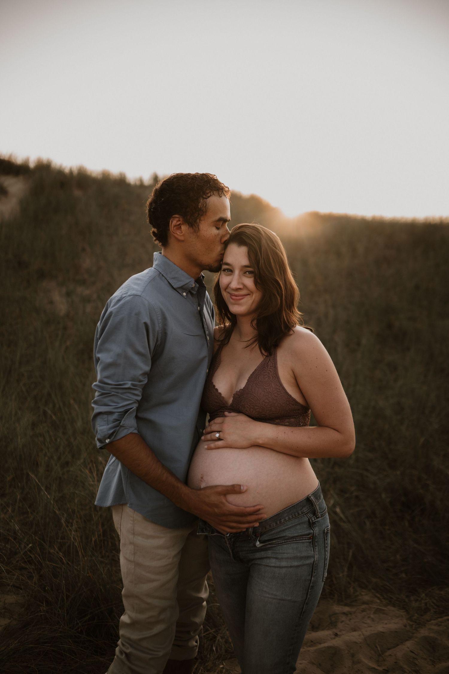 Intimate maternity photoshoot of expecting couple embracing at sunset on a hillside.