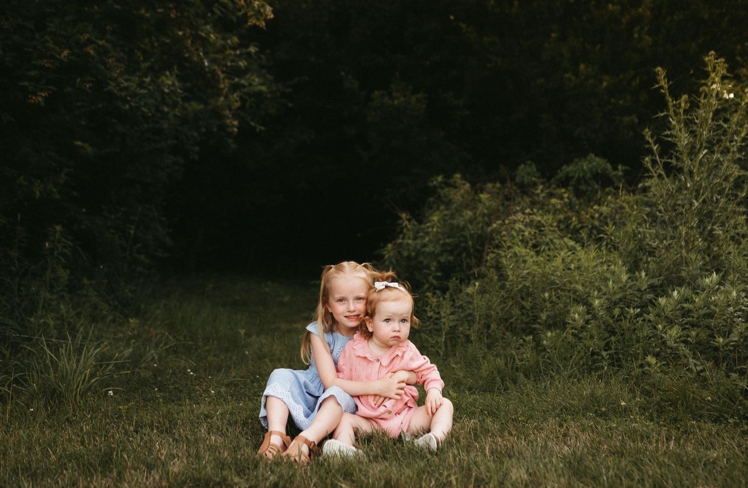 Two children sit together on green grass in front of a dark wooded background on a sunny day.