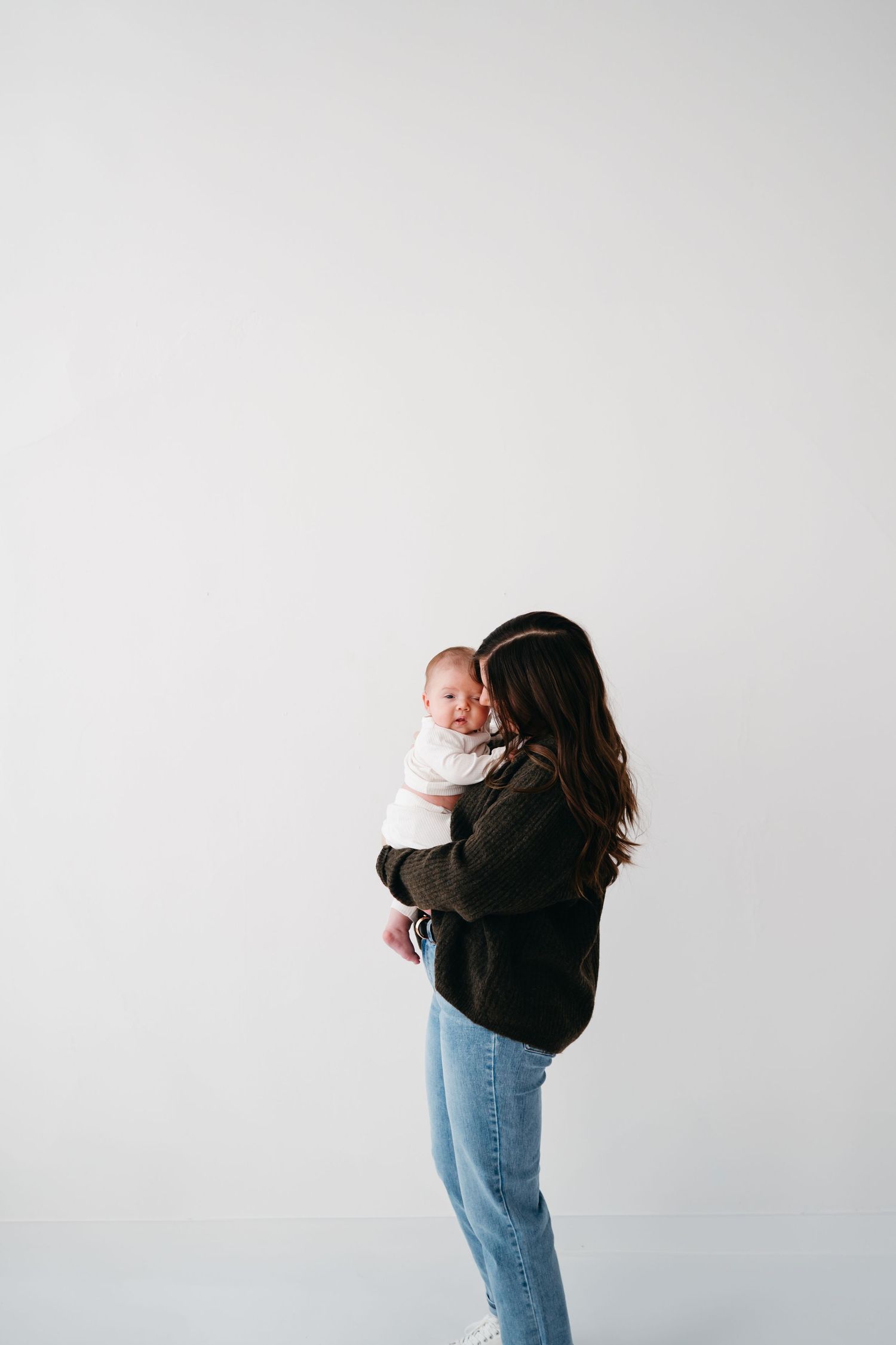 A parent holds and comforts their baby while standing against a minimalist white wall.